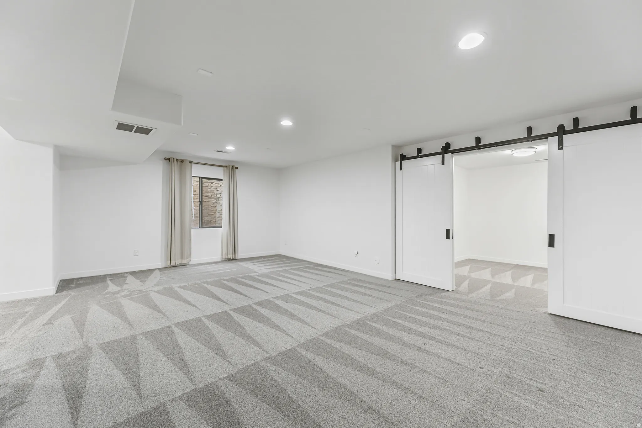 Spacious family room featuring patterned gray carpet flooring, recessed ceiling lighting, and a window with neutral curtains