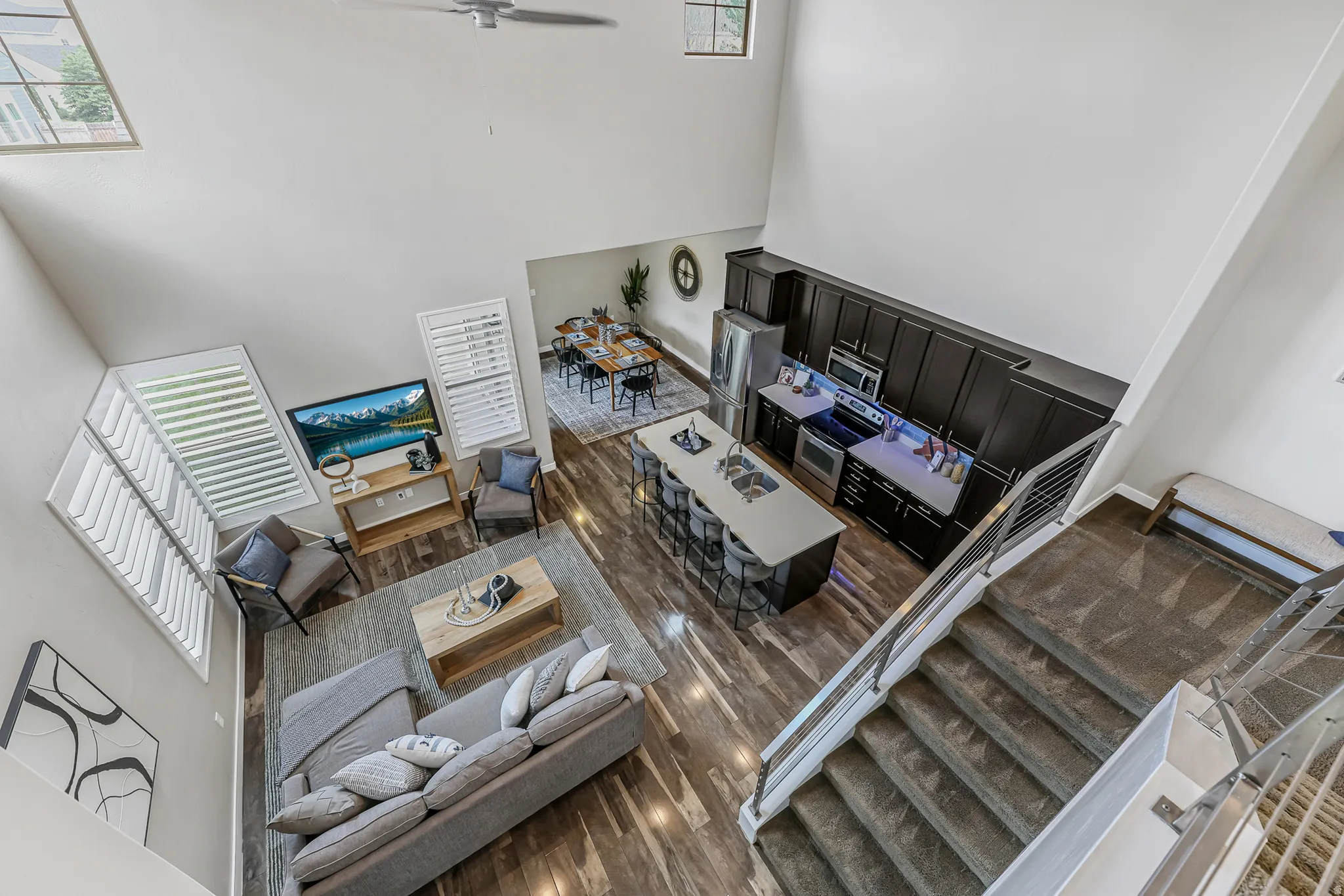 Open-concept living space featuring high ceilings, wood-finish flooring, a contemporary kitchen with dark cabinetry and stainless steel appliances, a kitchen island with seating, and a carpeted staircase with modern railings