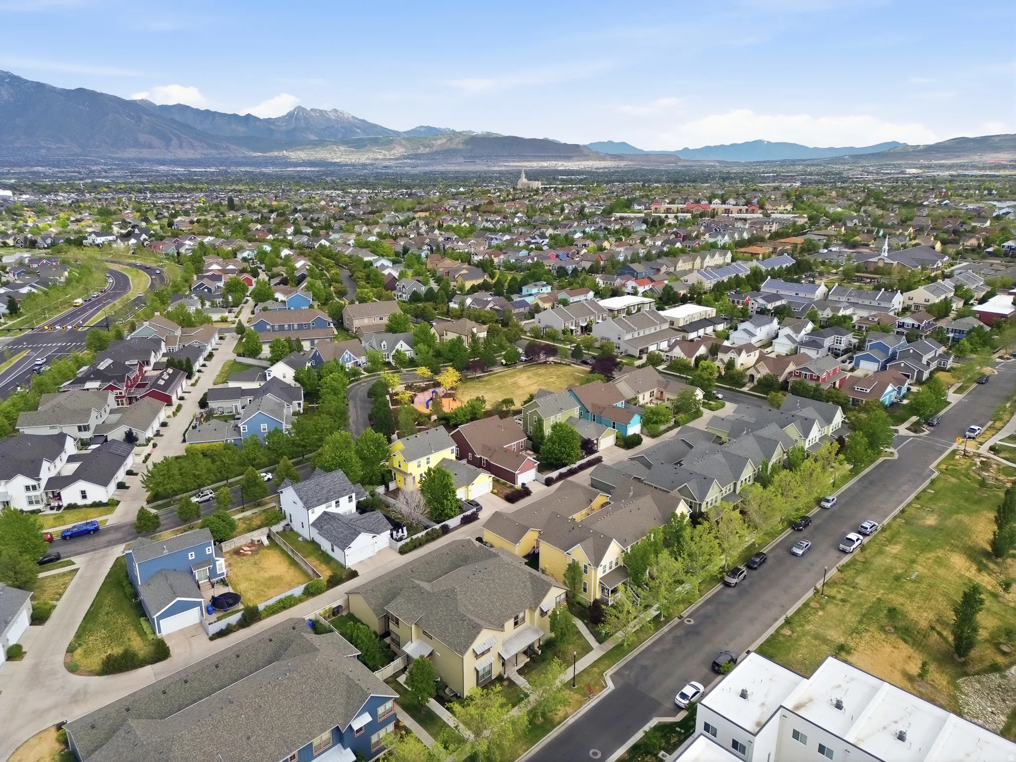 Expansive aerial perspective showcasing a residential neighborhood with varied architectural styles, mature trees, mountain range backdrop, paved streets, and a clear blue sky