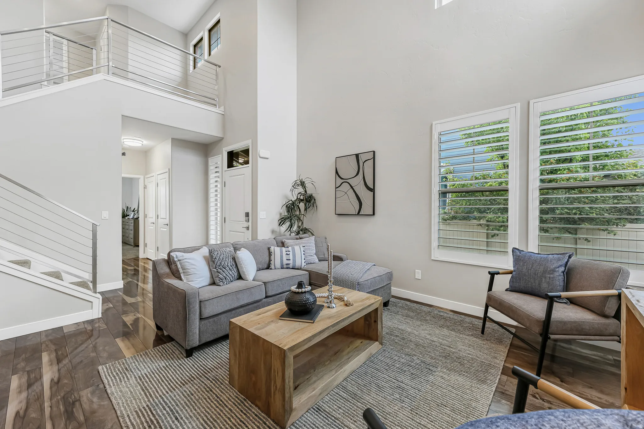 High ceiling living area featuring wood-finish flooring, large windows with plantation shutters, and a modern cable railing staircase