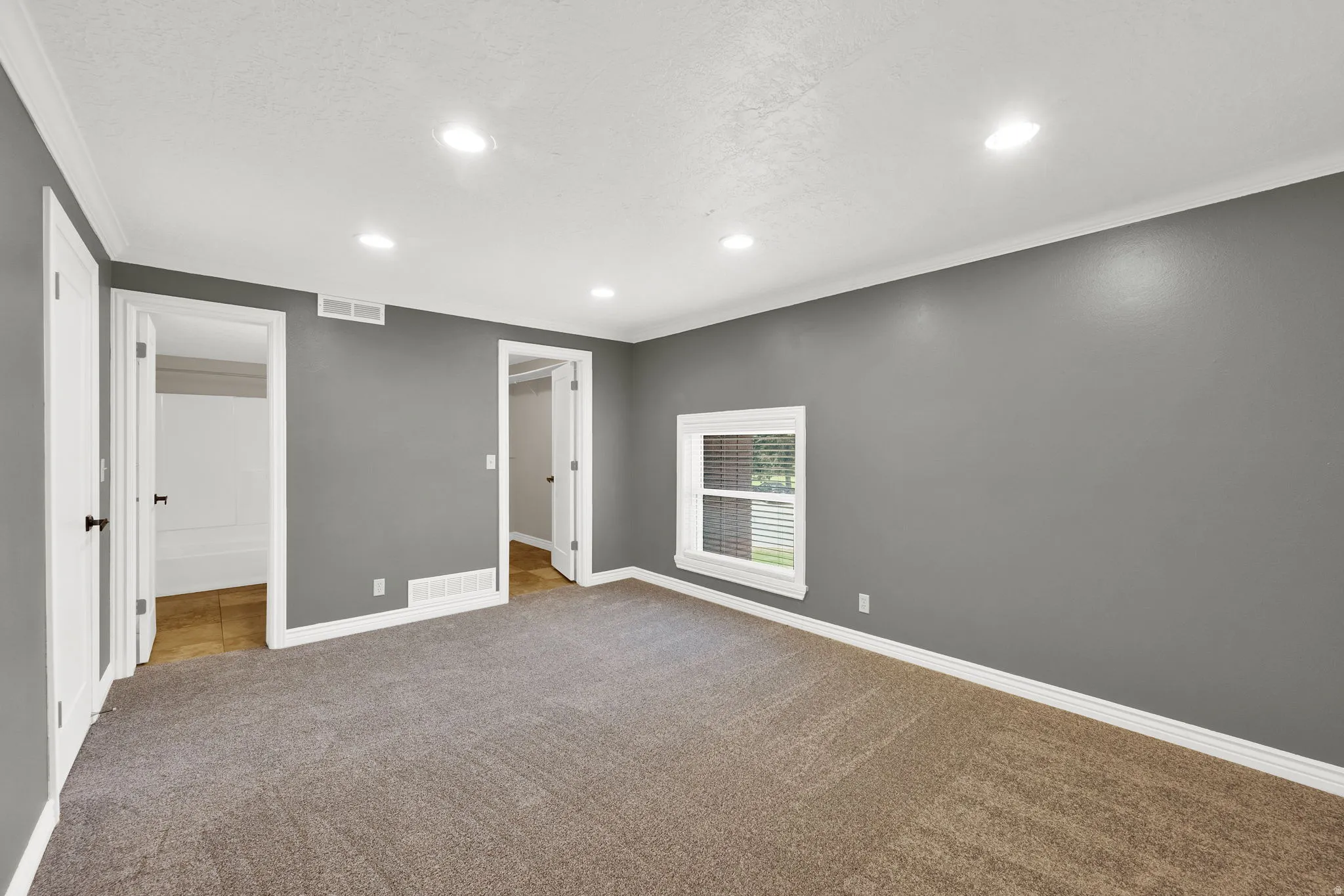 Spacious room with gray walls and neutral-toned carpeting, featuring recessed lighting, a single window, white trim, and two doorways