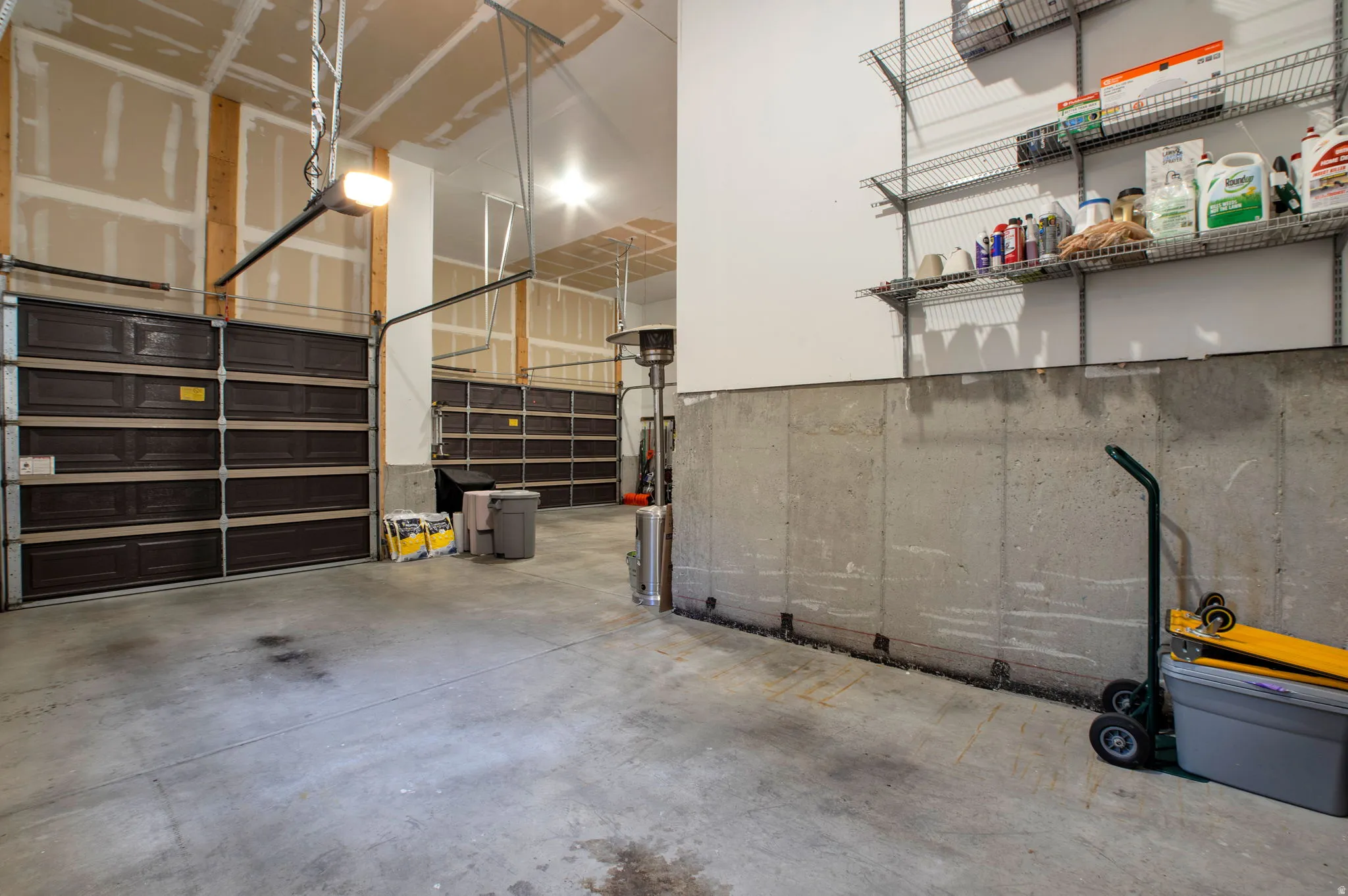 Two brown garage doors, concrete flooring, exposed wood framing, concrete foundation wall, and wall-mounted wire shelving