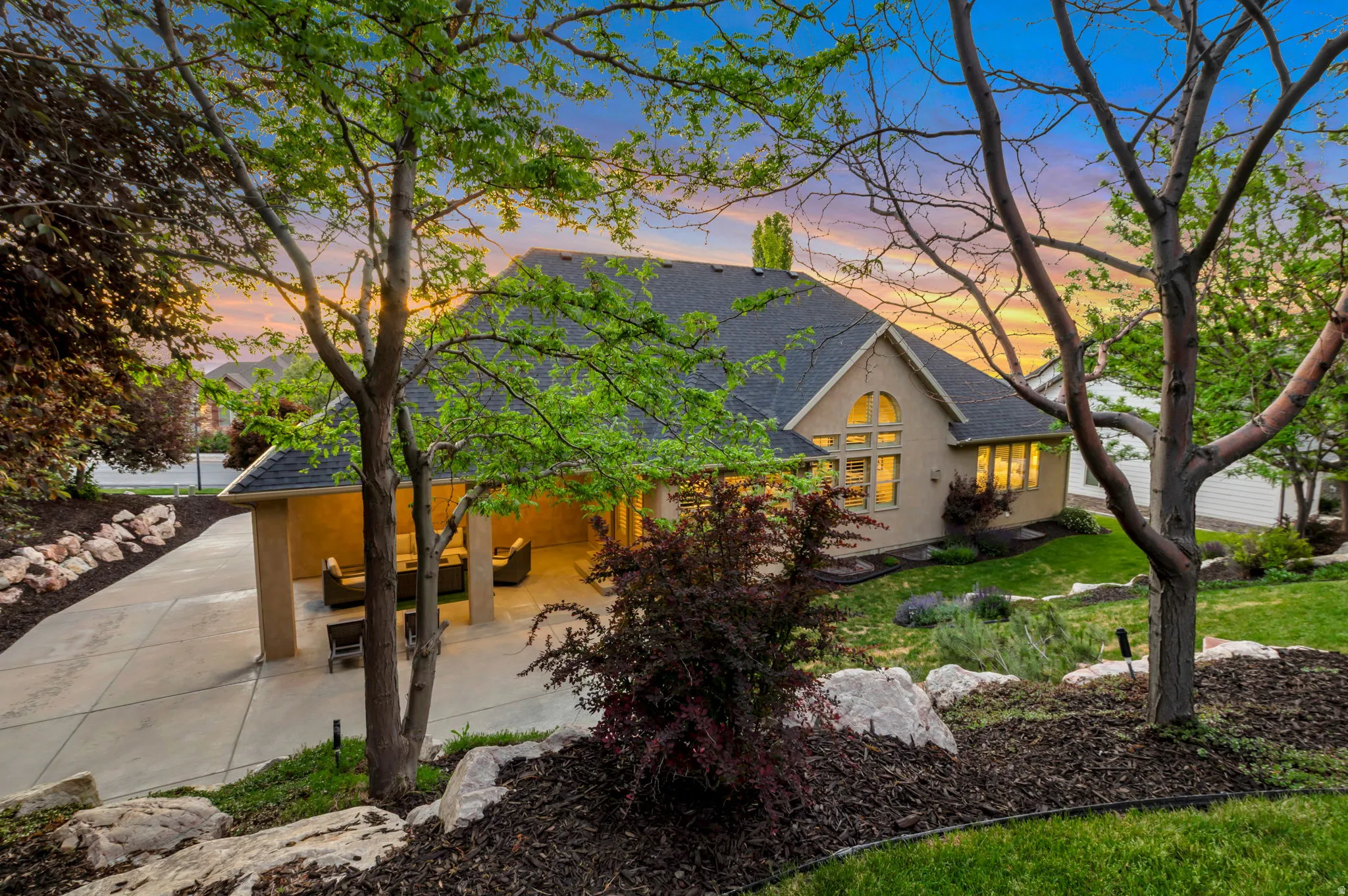 Expansive rear exterior featuring a covered patio, broad concrete driveway, and mature trees