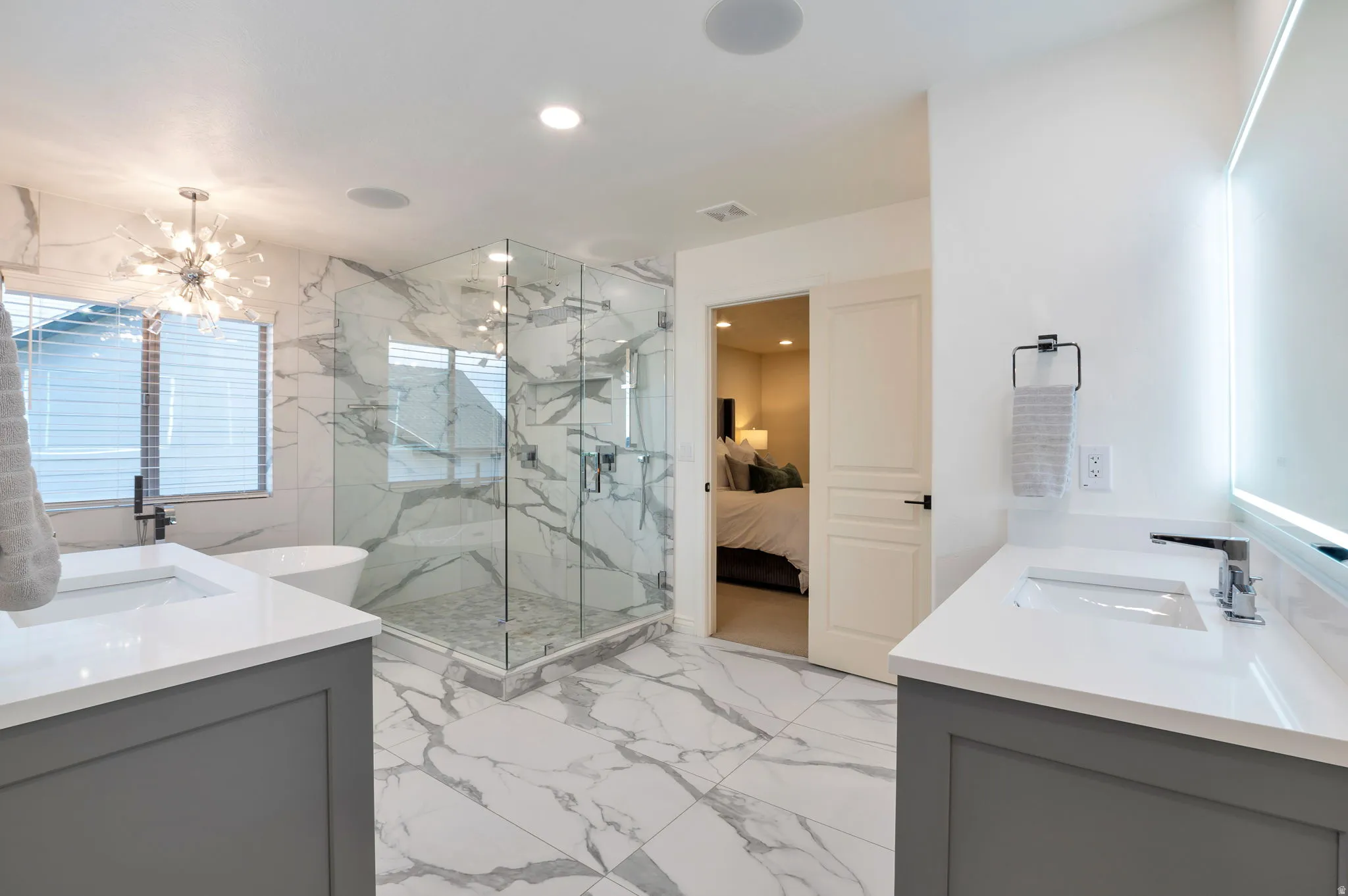 Spacious bathroom featuring large format marble-look tile flooring and walls, a glass-enclosed shower, dual vanities with white countertops, and a freestanding soaking tub