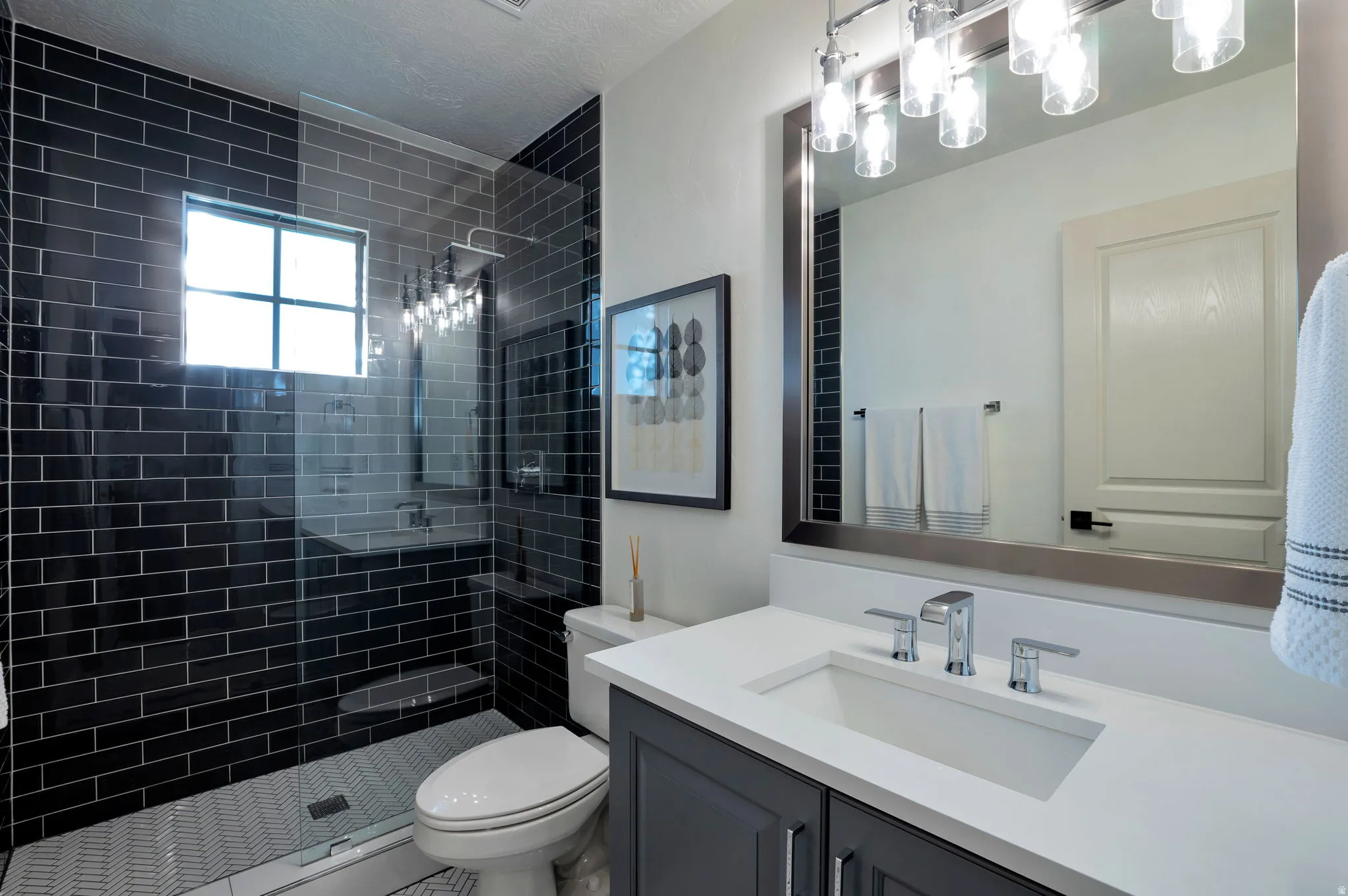Bathroom featuring a frameless glass shower enclosure with dark subway tile, a white vanity with an integrated sink, a rectangular framed mirror, and a contemporary multi-bulb light fixture