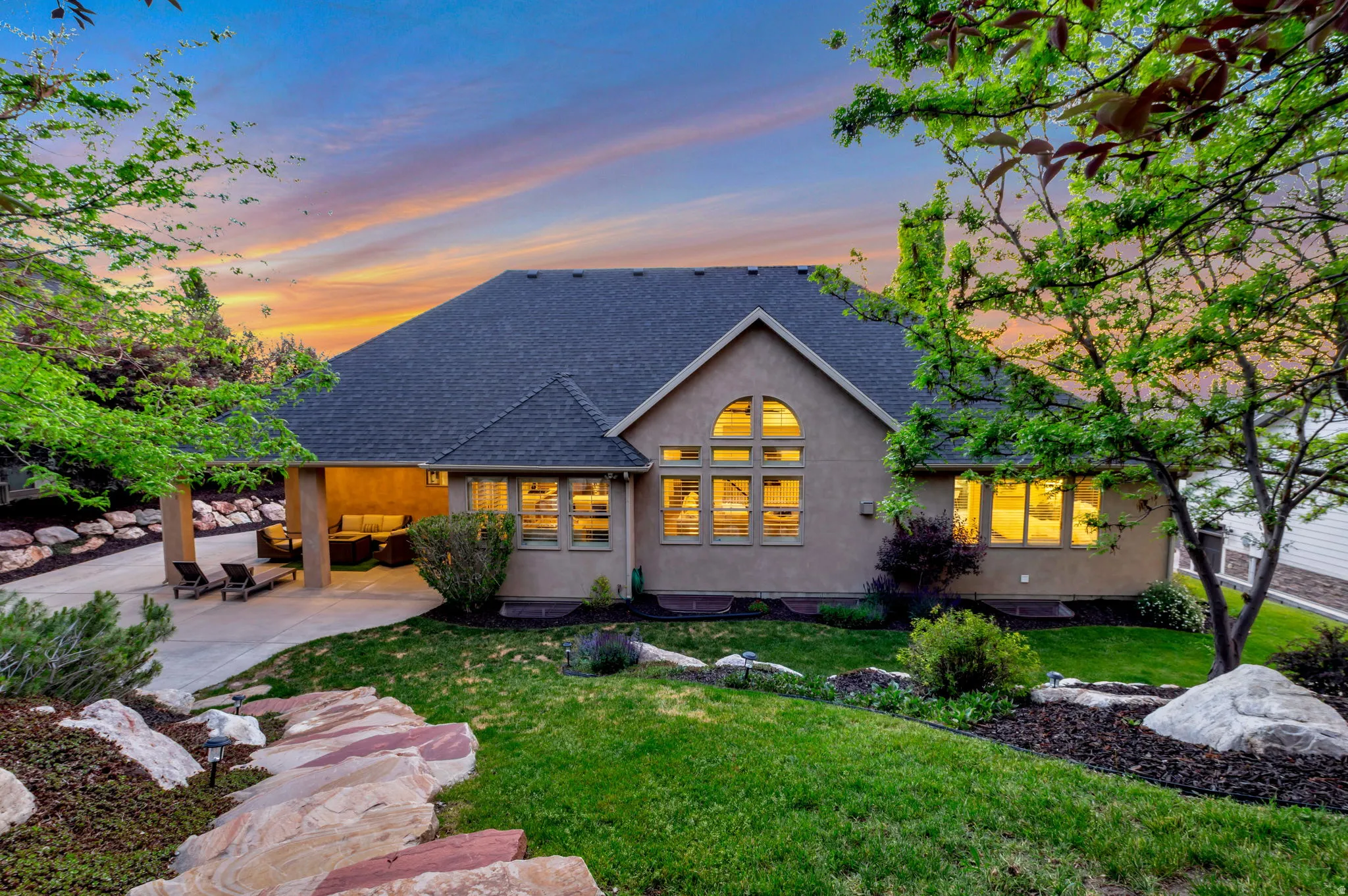 Rear exterior featuring a covered patio, extensive backyard landscaping with natural rock elements, and a multi-pane arched window