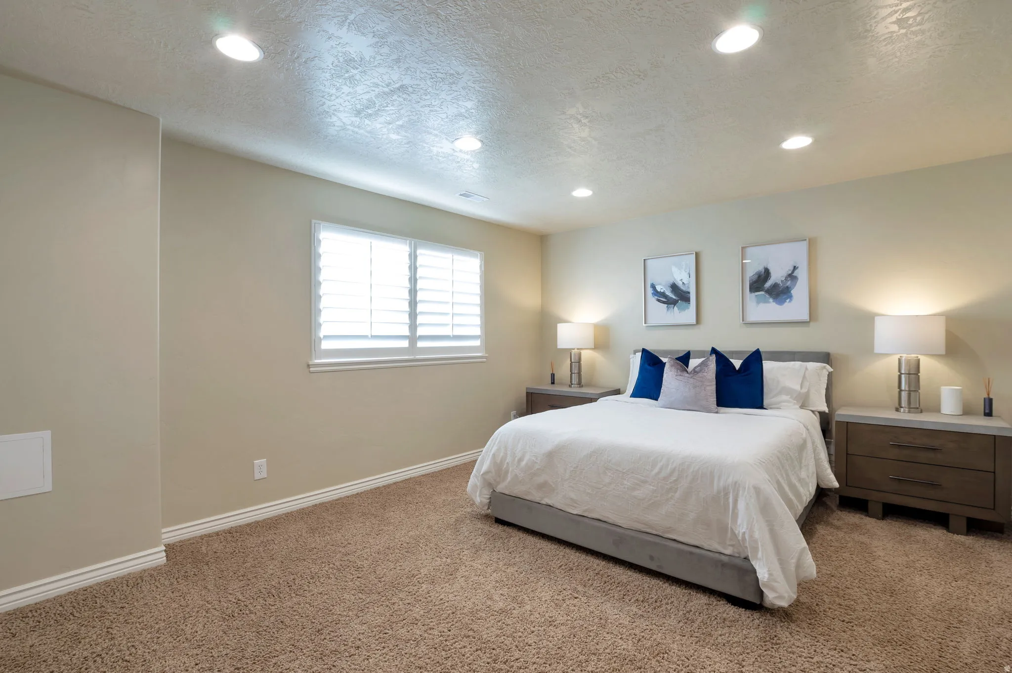 Recessed ceiling lighting illuminates a spacious room featuring textured carpet flooring, neutral-toned walls, and a window with white plantation shutters