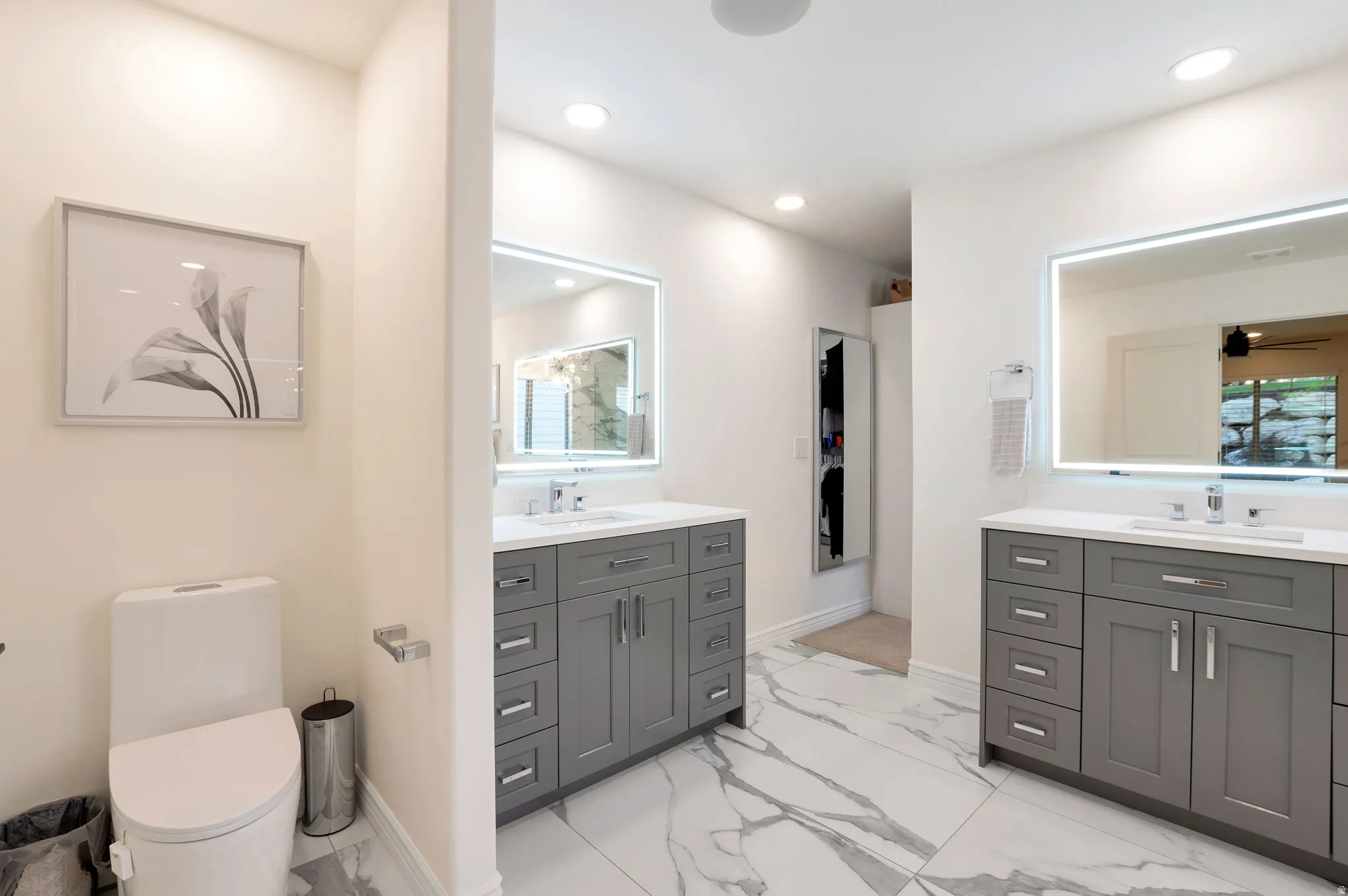 Spacious bathroom featuring dual gray vanities with white countertops, illuminated mirrors, recessed lighting, and large format marble-look tile flooring