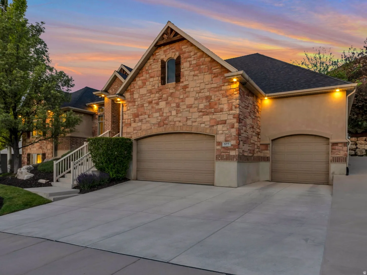 Stone and stucco exterior featuring multiple garages, a concrete driveway, and a tiered landscaped yard with mature trees