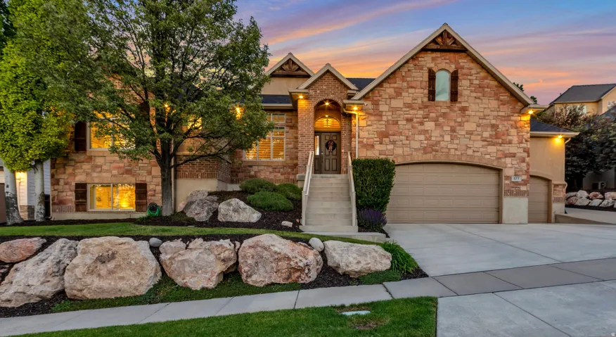 Stone and brick facade featuring an arched entryway with a wooden door, a multi-car garage with arched doors, mature trees, and a landscaped rock garden