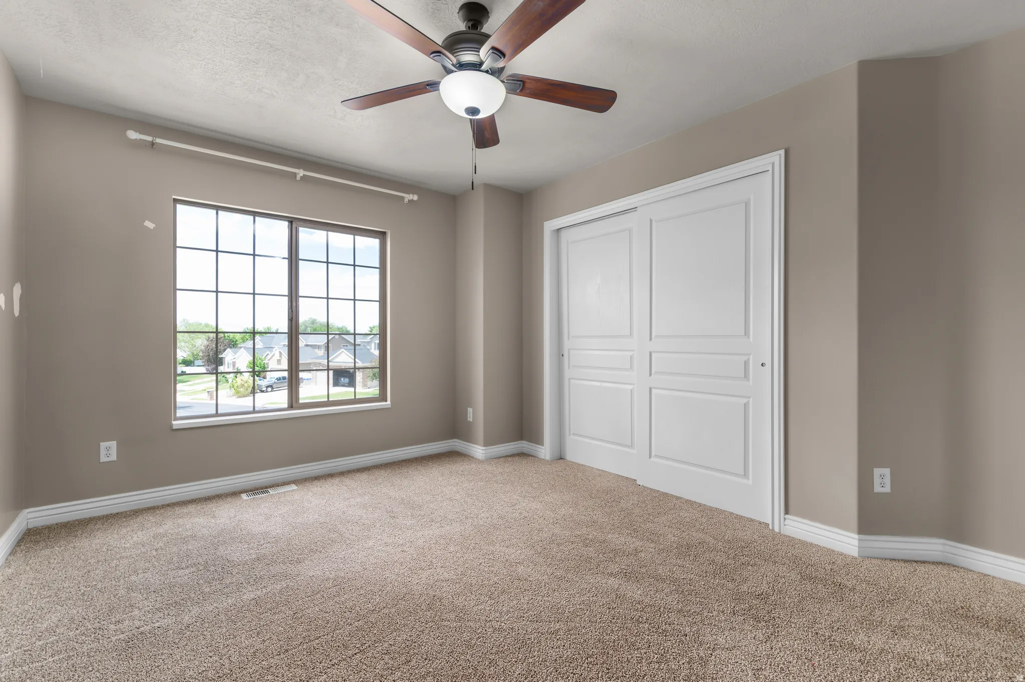Spacious room featuring neutral carpeting, a large window with grid pattern, a ceiling fan with integrated lighting, and white bi-fold closet doors