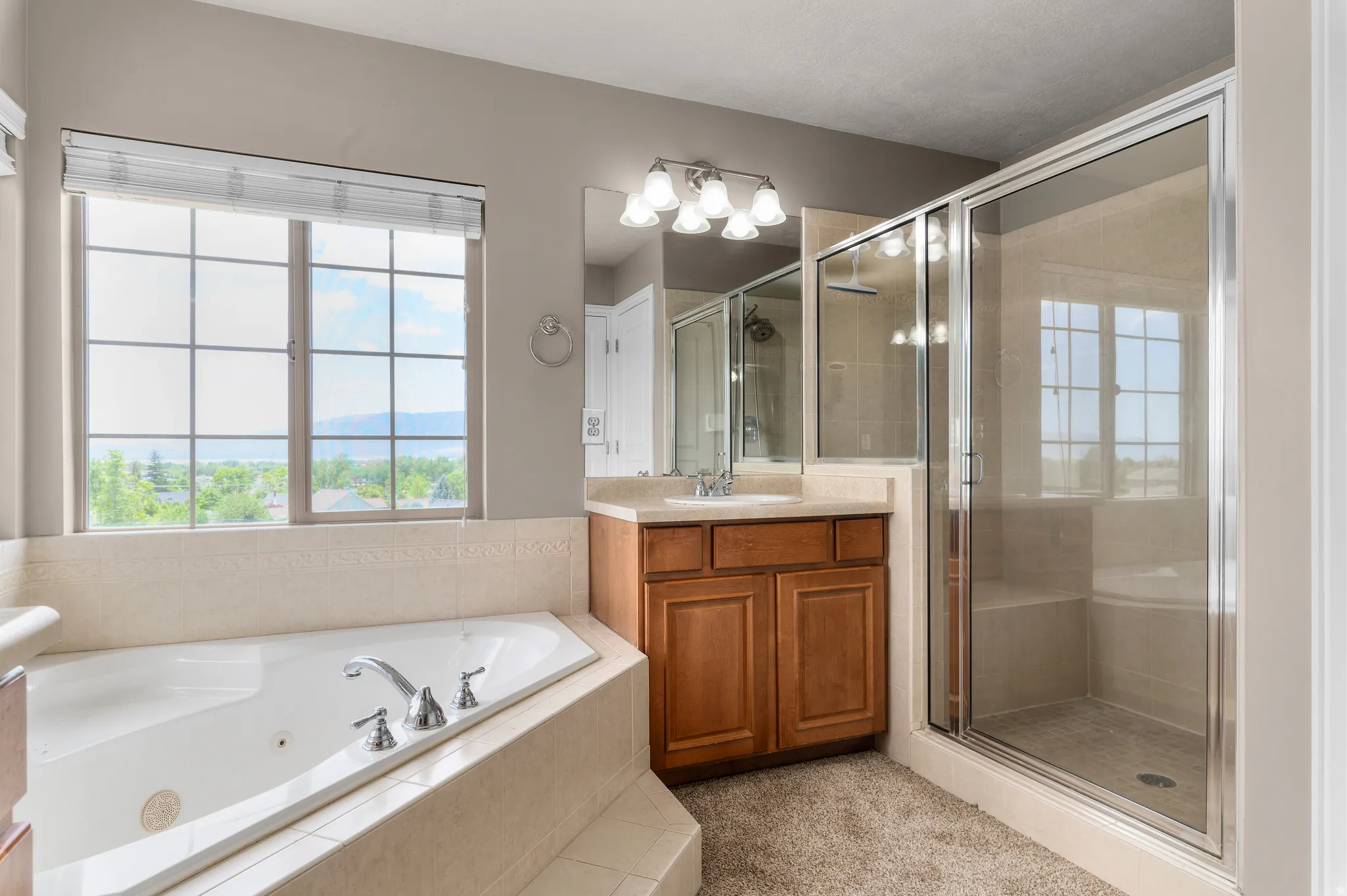 Bathroom featuring a jetted soaking tub with tile surround, a separate glass-enclosed shower with tiled walls and built-in bench, a single vanity with wood cabinetry and a light-toned countertop, and grid-pattern windows with roller shades