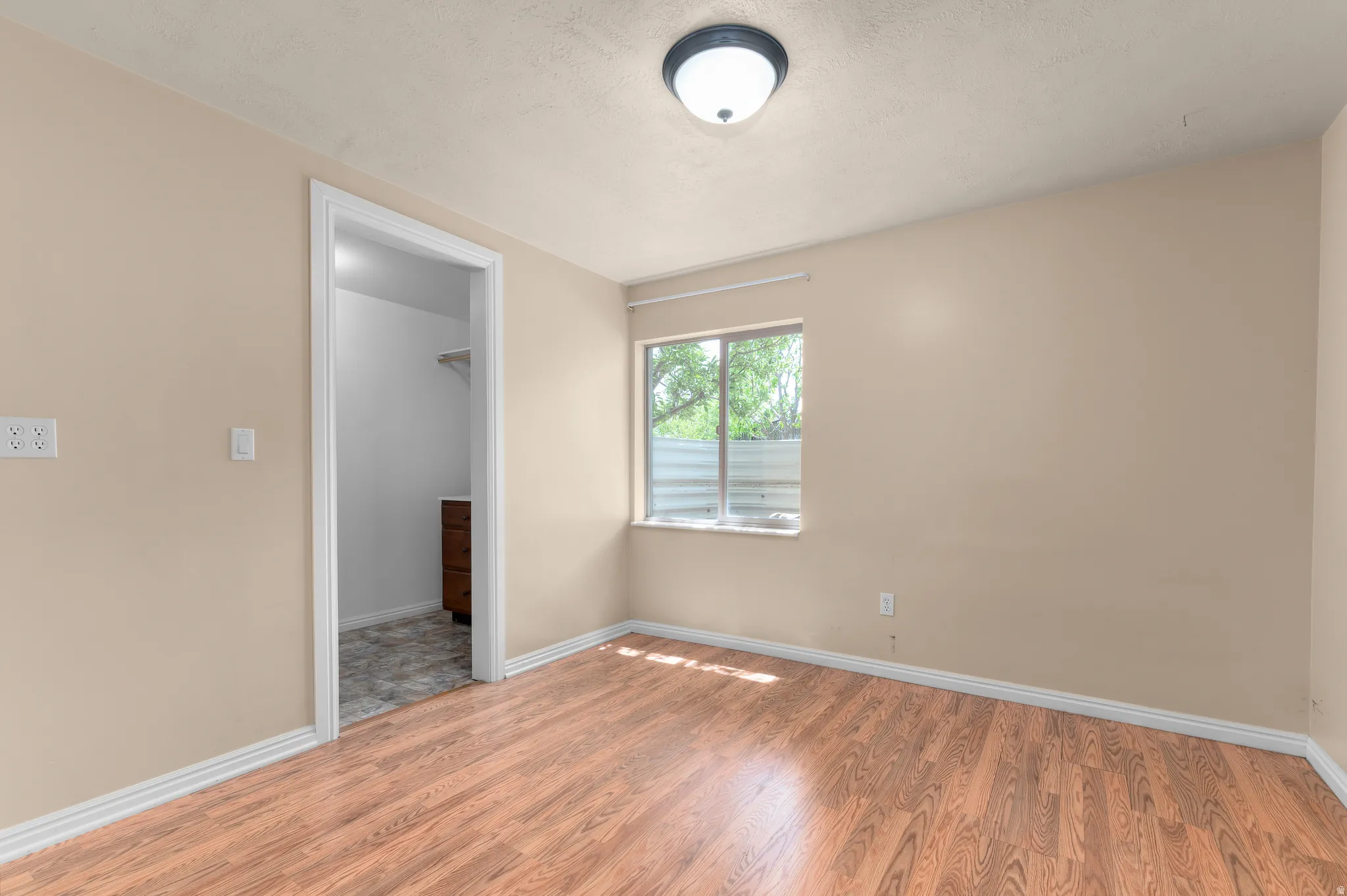 Interior room featuring wood-finish flooring, light beige wall paint, white baseboards, and a window with a slatted privacy screen