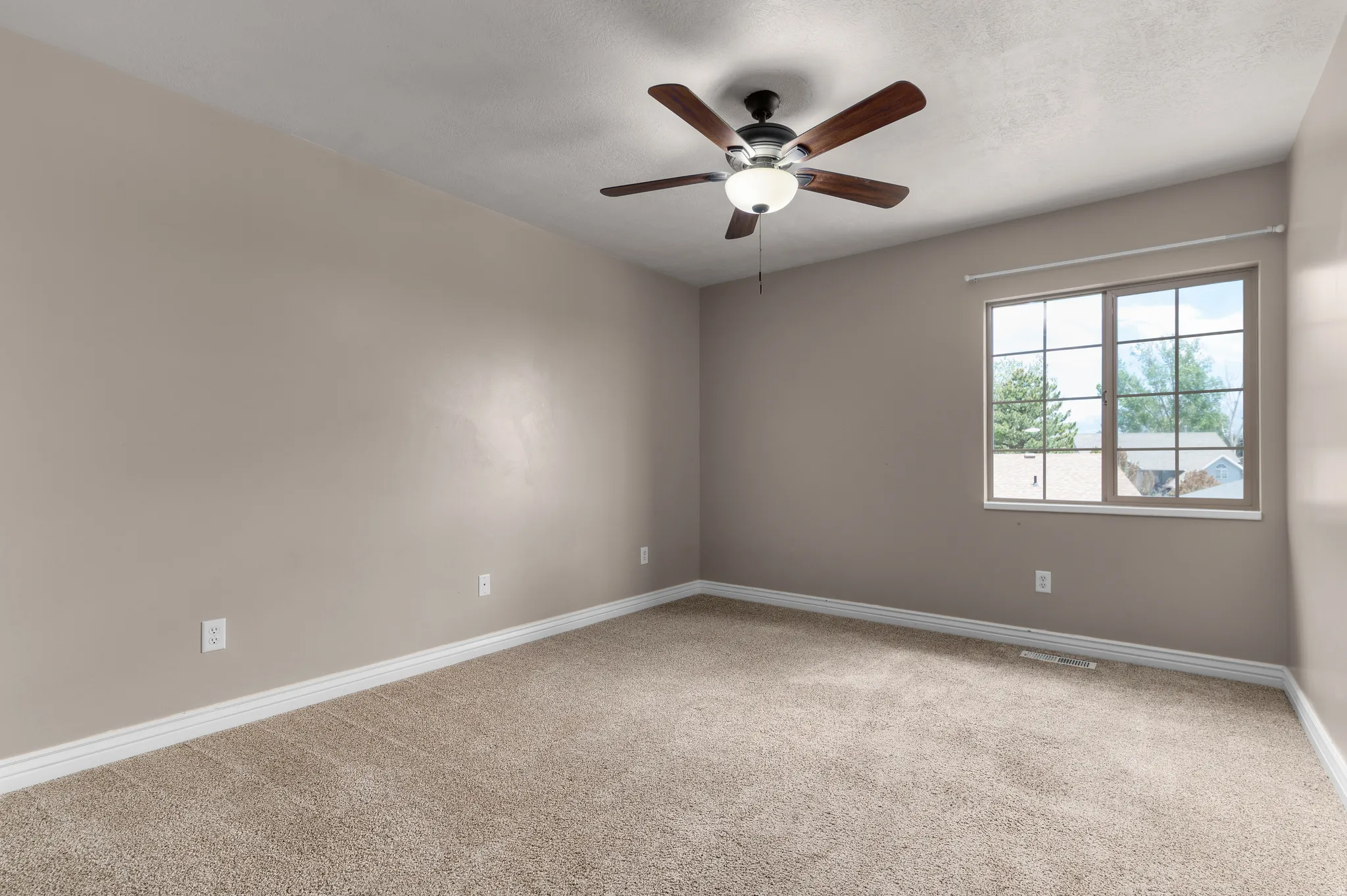 Neutral-toned room featuring a large window with grid panes, a ceiling fan with integrated lighting, textured carpet flooring, and white baseboards