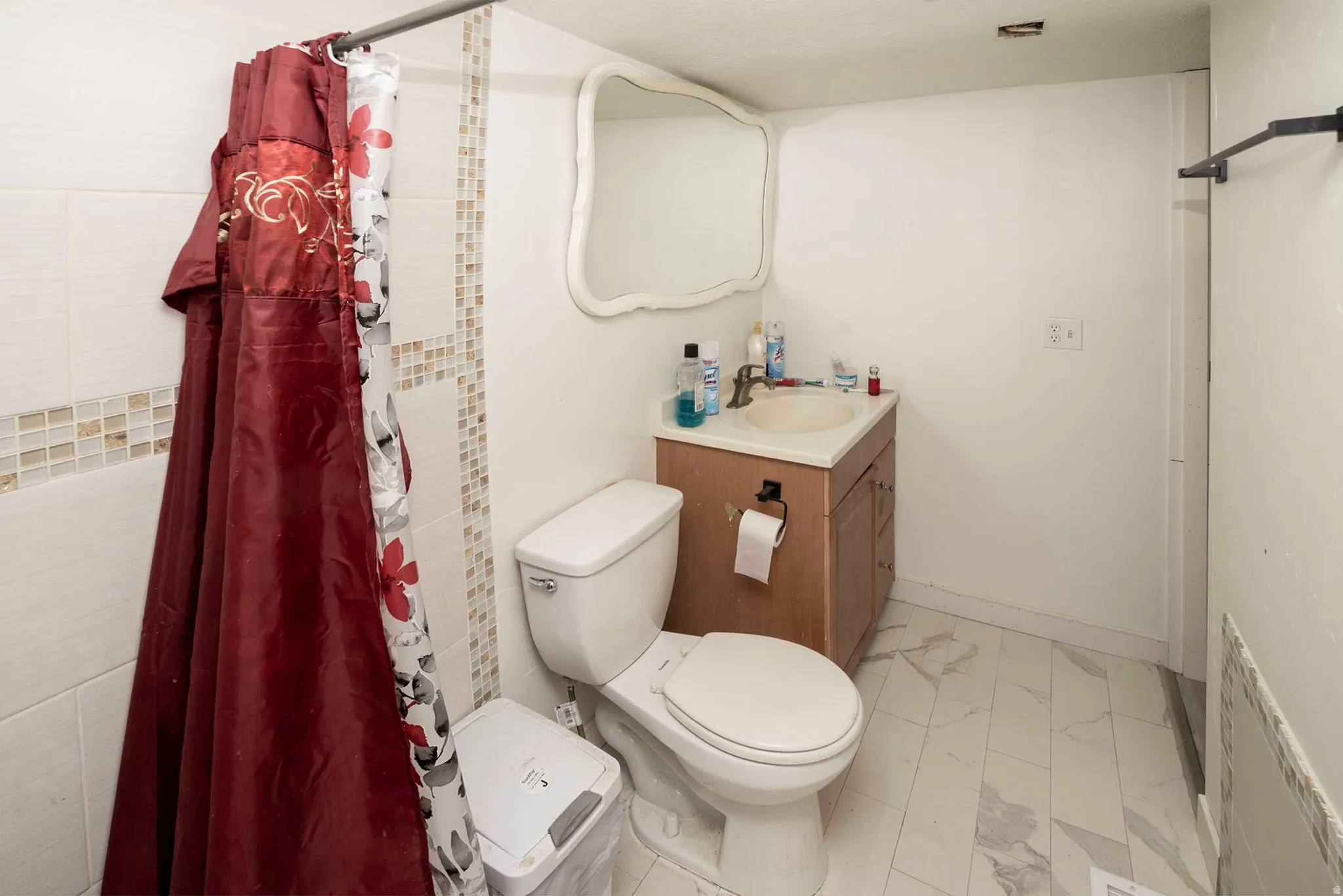 Bathroom featuring a wood-finish vanity with an integrated sink, a commode, a shower area with white tile and mosaic accents, and light-toned tile flooring