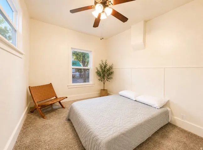 Unit 2 bedroom carpeted featuring a ceiling fan with integrated lighting, two windows with white trim, and a wall with wainscoting detail