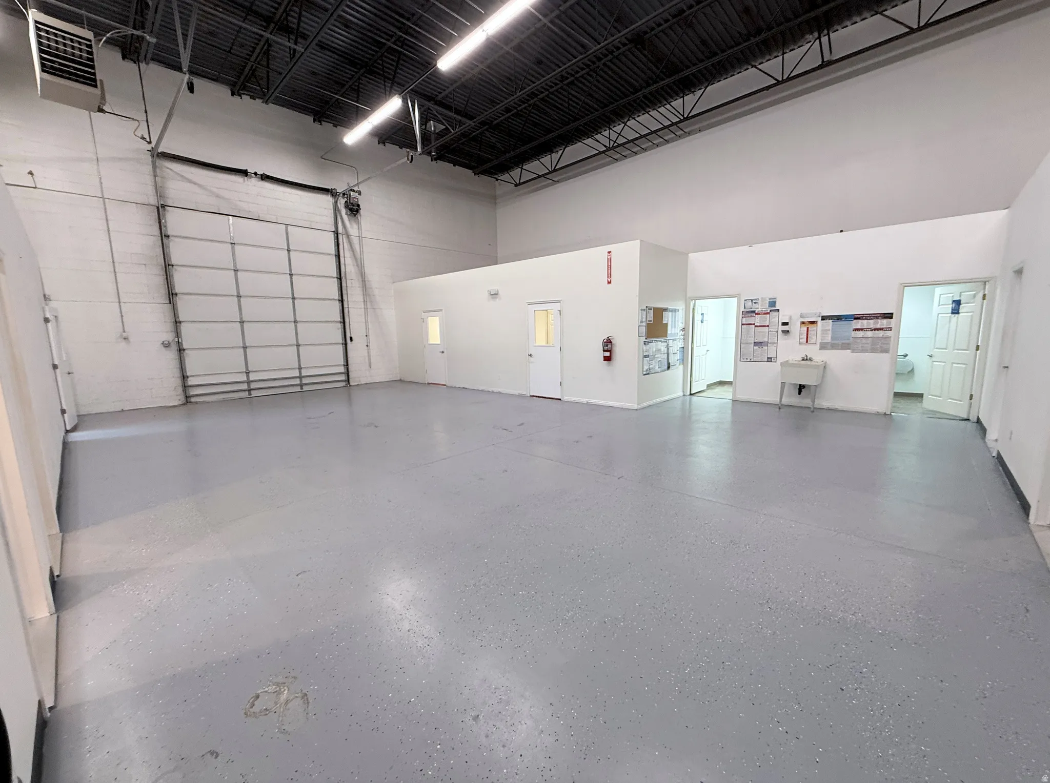 Spacious commercial interior featuring a large roll-up door, high ceilings with exposed ductwork, gray speckled flooring, white painted walls, and multiple interior access doors