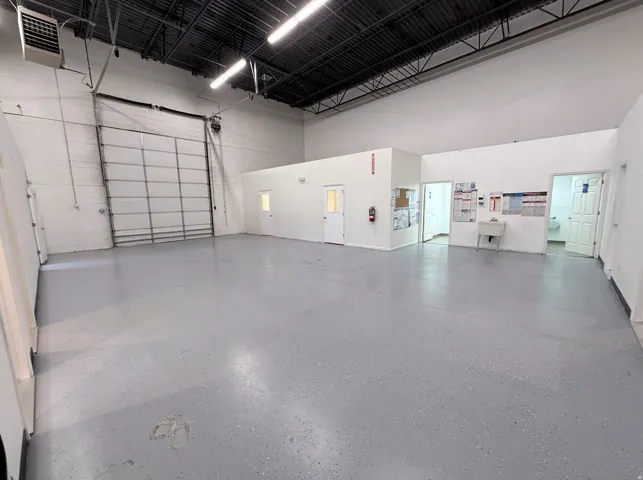 Spacious commercial interior featuring a large roll-up door, high ceilings with exposed ductwork, gray speckled flooring, white painted walls, and multiple interior access doors