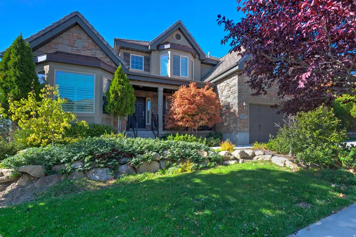 Stately residence featuring a stone and stucco exterior, prominent bay windows, a covered entry porch, an integrated two-car garage, and mature landscaping with a rock retaining wall