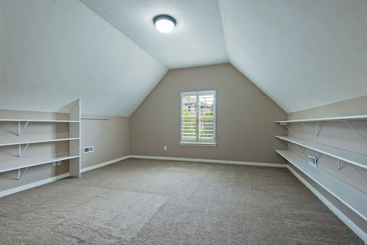 Expansive room featuring sloped ceilings, plush carpet, and a window with white plantation shutters