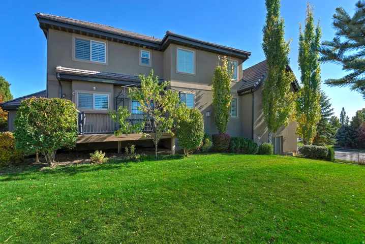 Expansive green lawn bordering a multi-level structure with a stucco exterior, multiple windows featuring louvered shutters, and a rear deck with railing