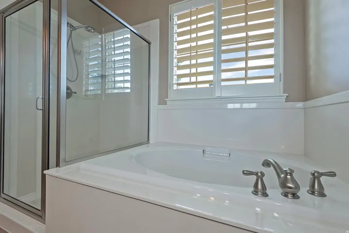 Bathtub with brushed nickel fixtures, plantation shutters, and a glass-enclosed shower