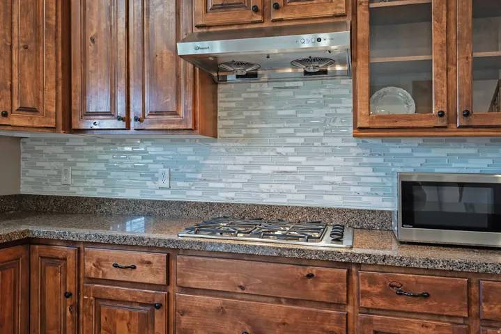 Kitchen featuring wood cabinetry, a glass tile backsplash, stone-finish countertops, a stainless steel gas cooktop, and a stainless steel range hood