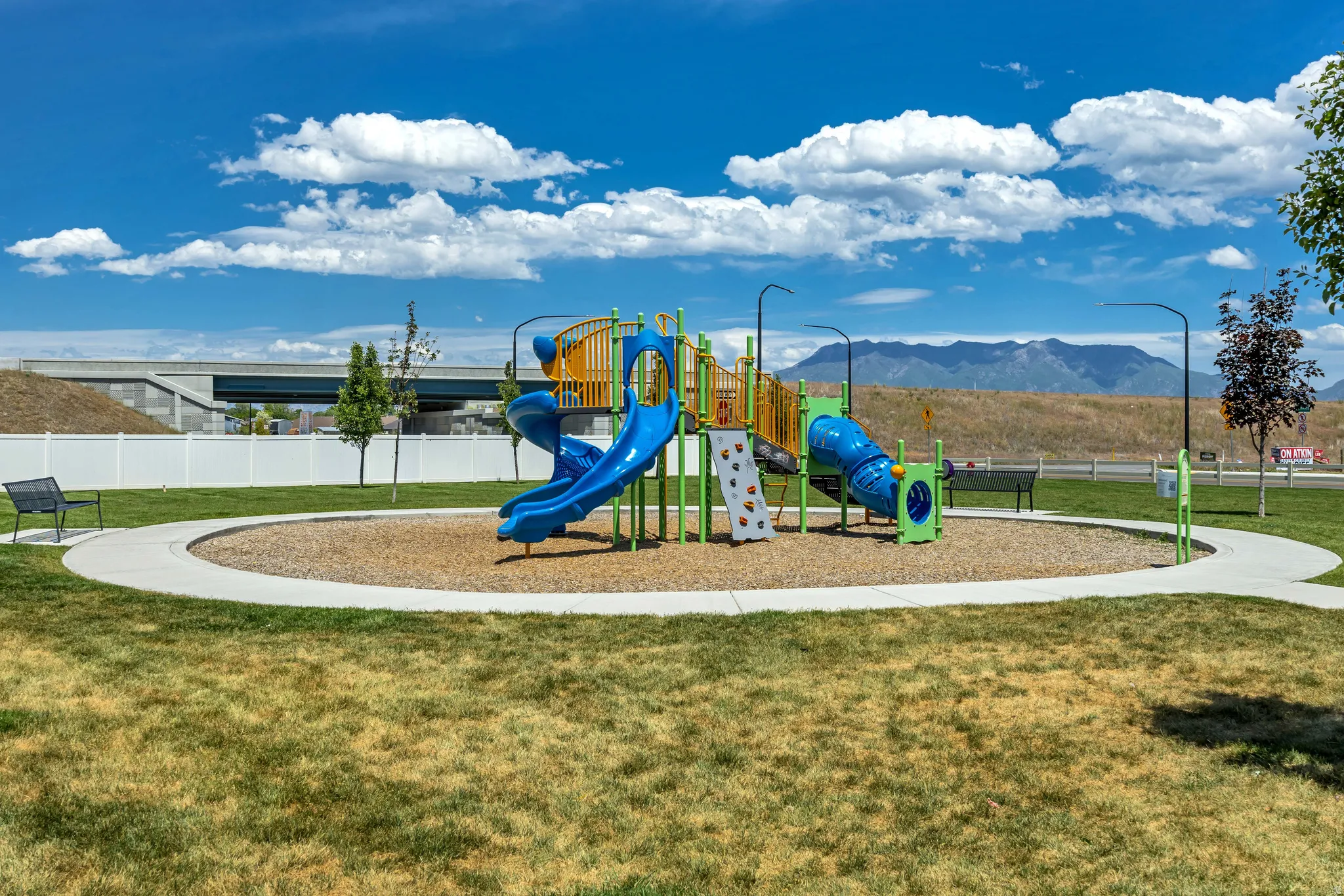 Community playground featuring a multi-level play structure with slides and climbing walls, surrounded by a wood chip surface and a concrete walking path