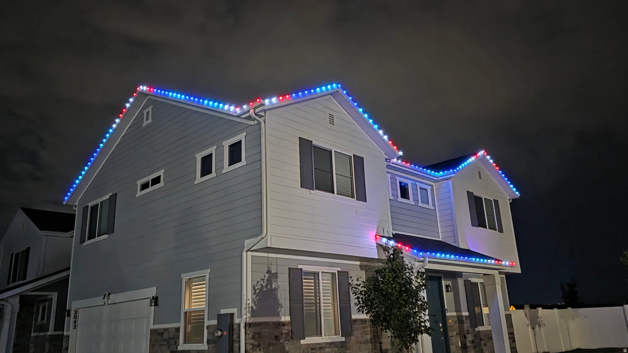 Two-story residence featuring light-toned siding and dark shutters-permanent exterior custom holiday lighting