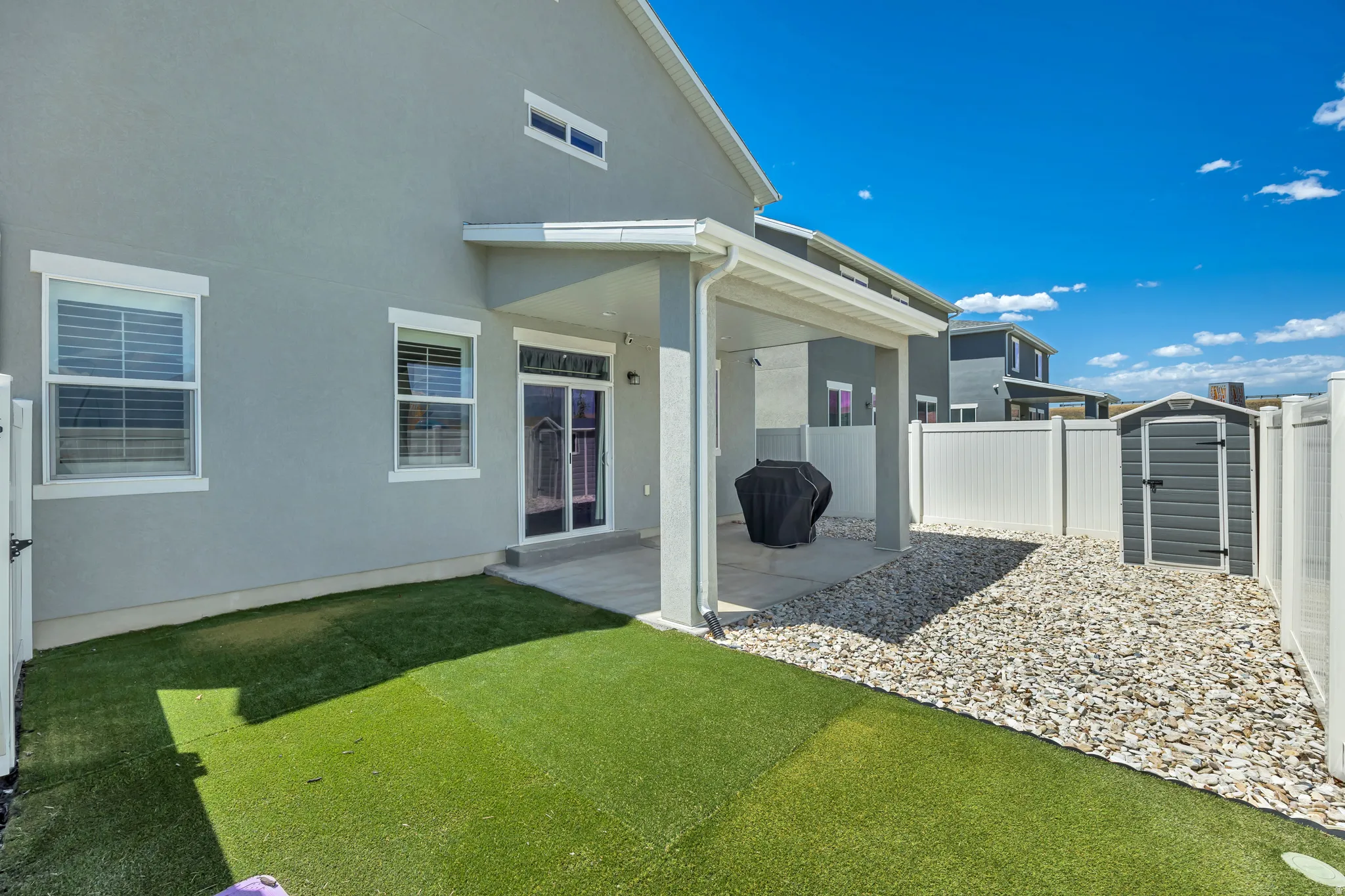 Rear exterior featuring a covered patio, artificial turf, gravel landscaping, a storage shed, and white privacy fencing