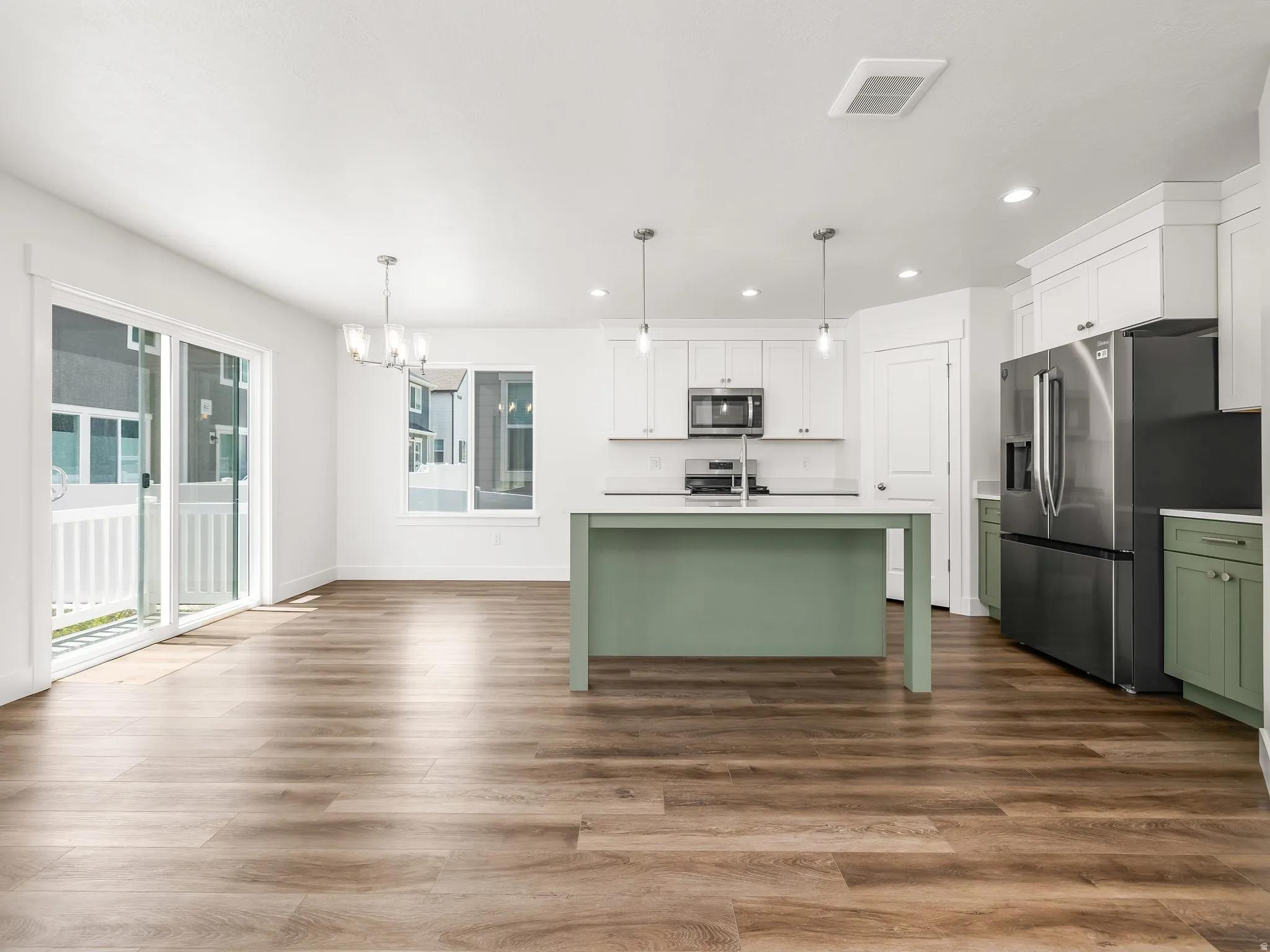 Open-concept kitchen and dining area featuring wood-finish flooring, a contemporary kitchen island with a light green finish, white cabinetry, stainless steel appliances, and a sliding glass door leading to a balcony