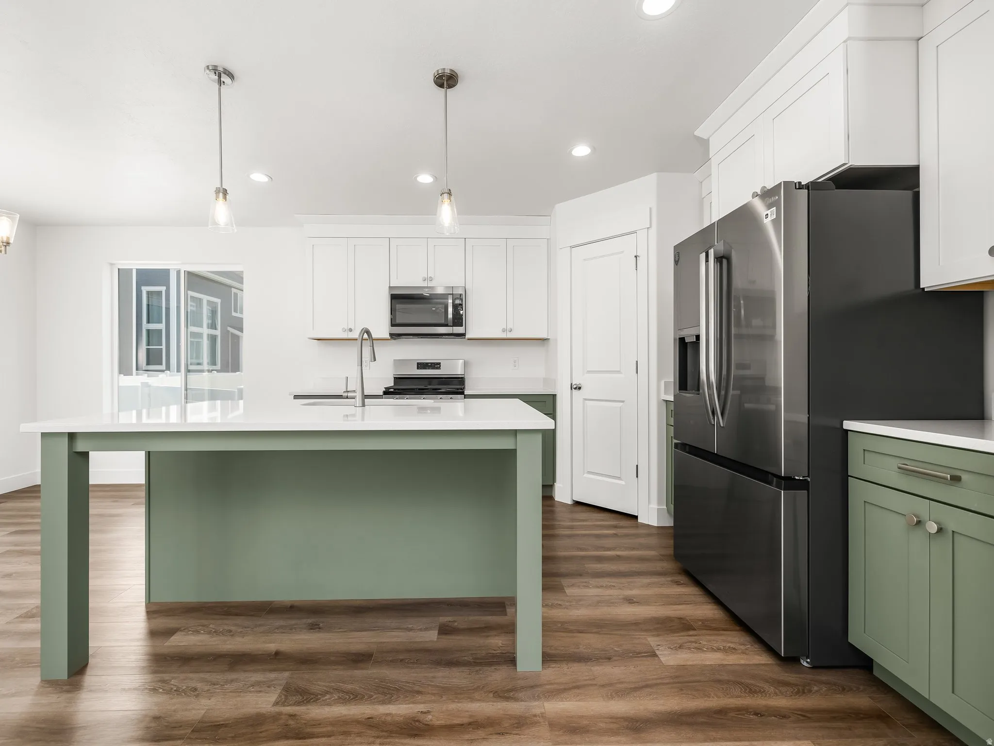 Modern kitchen featuring a central island with a light green base, white countertops, stainless steel appliances, white upper cabinetry, and wood-finish flooring