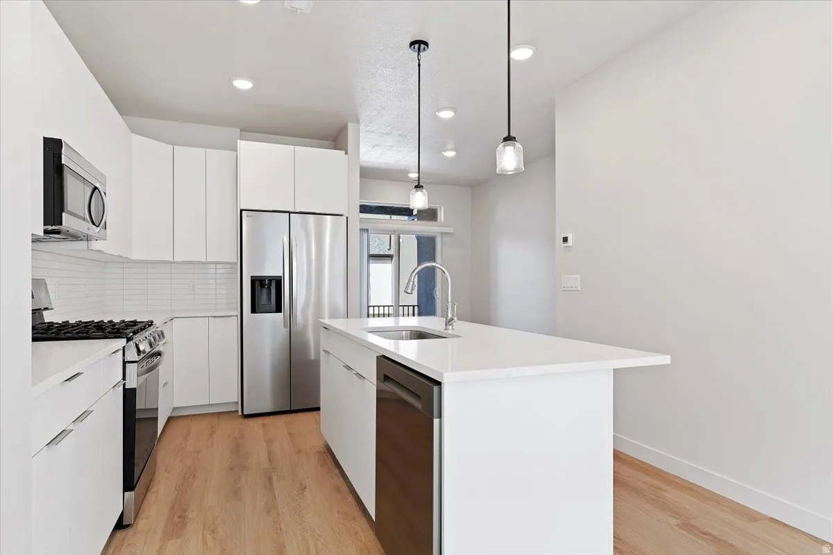 Modern kitchen featuring a central island with integrated sink, stainless steel appliances, white flat-panel cabinetry, and wood-finish flooring