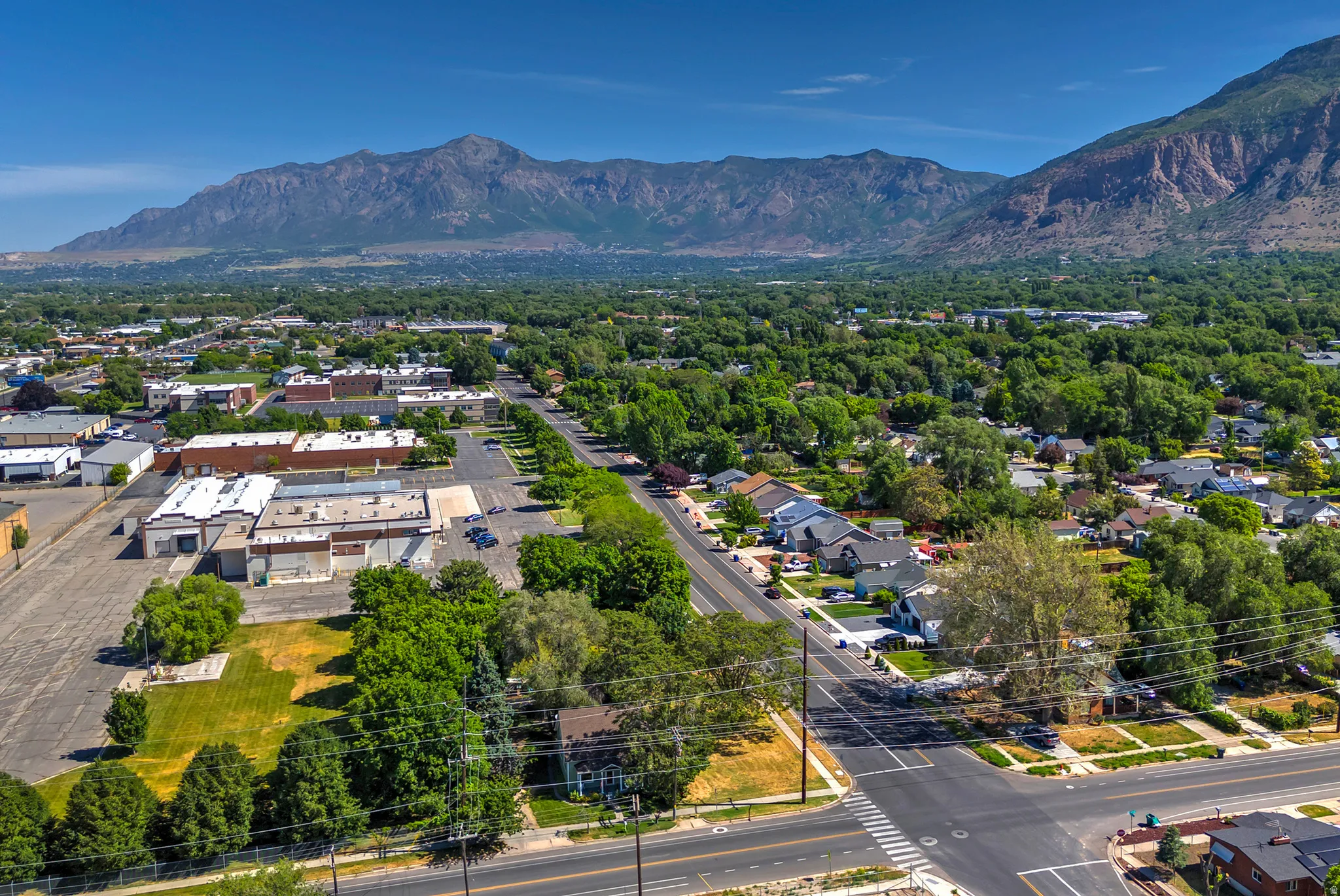 Expansive aerial perspective showcasing a residential neighborhood with tree-lined streets, a commercial area, and a prominent mountain range backdrop