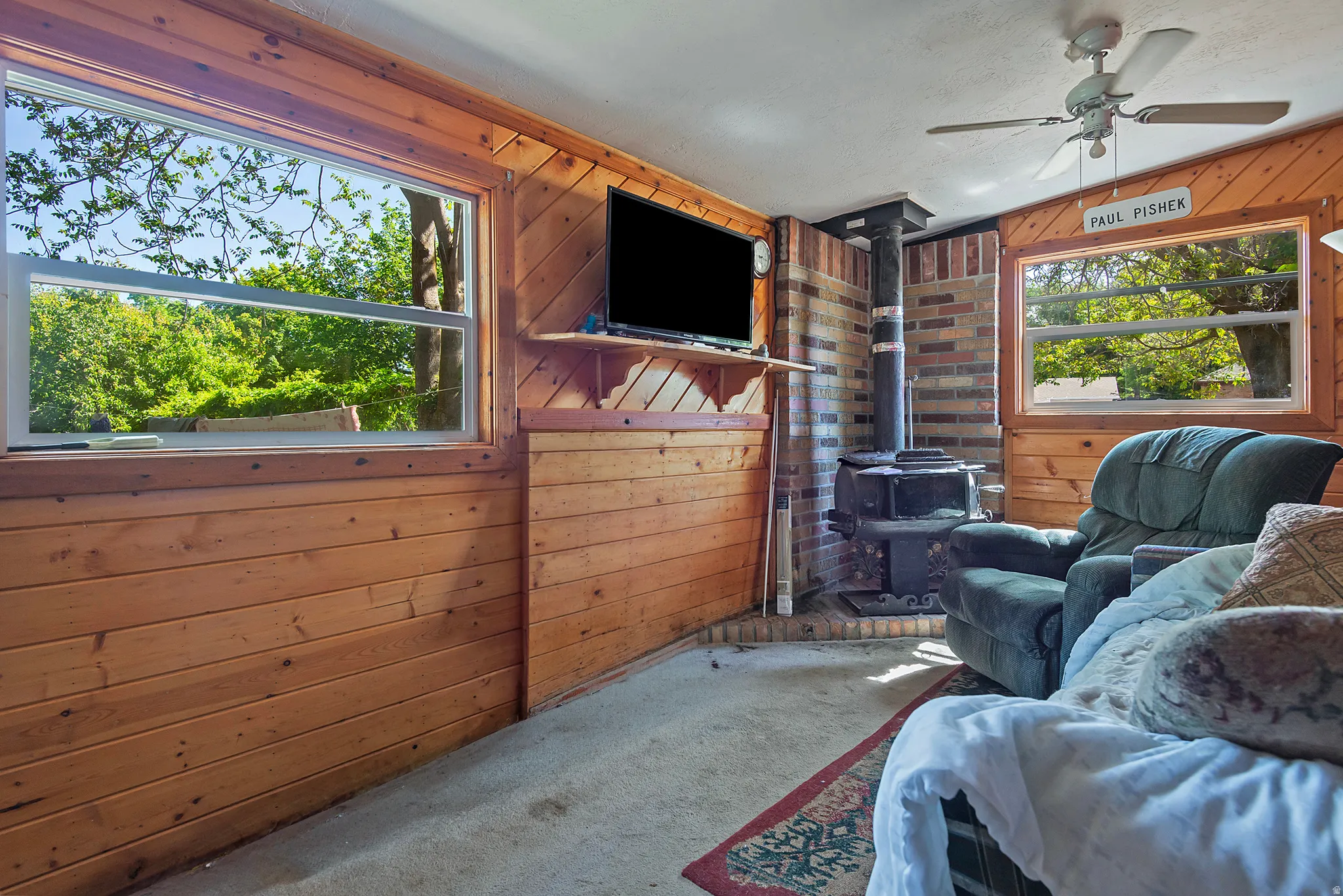 Room featuring wood paneling, a ceiling fan, and a freestanding wood-burning stove with a brick surround