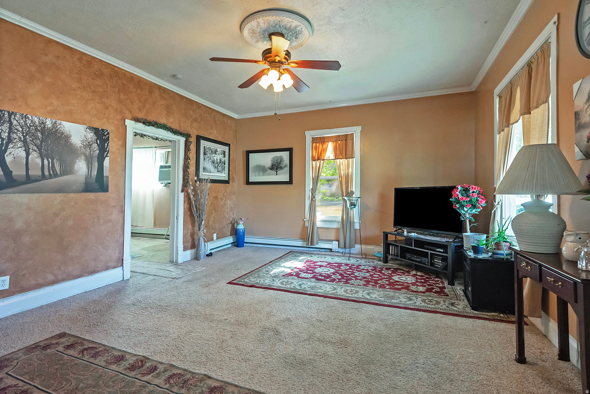 Spacious living area featuring neutral carpeting, warm wall tones, white crown molding, and a ceiling fan with integrated lighting