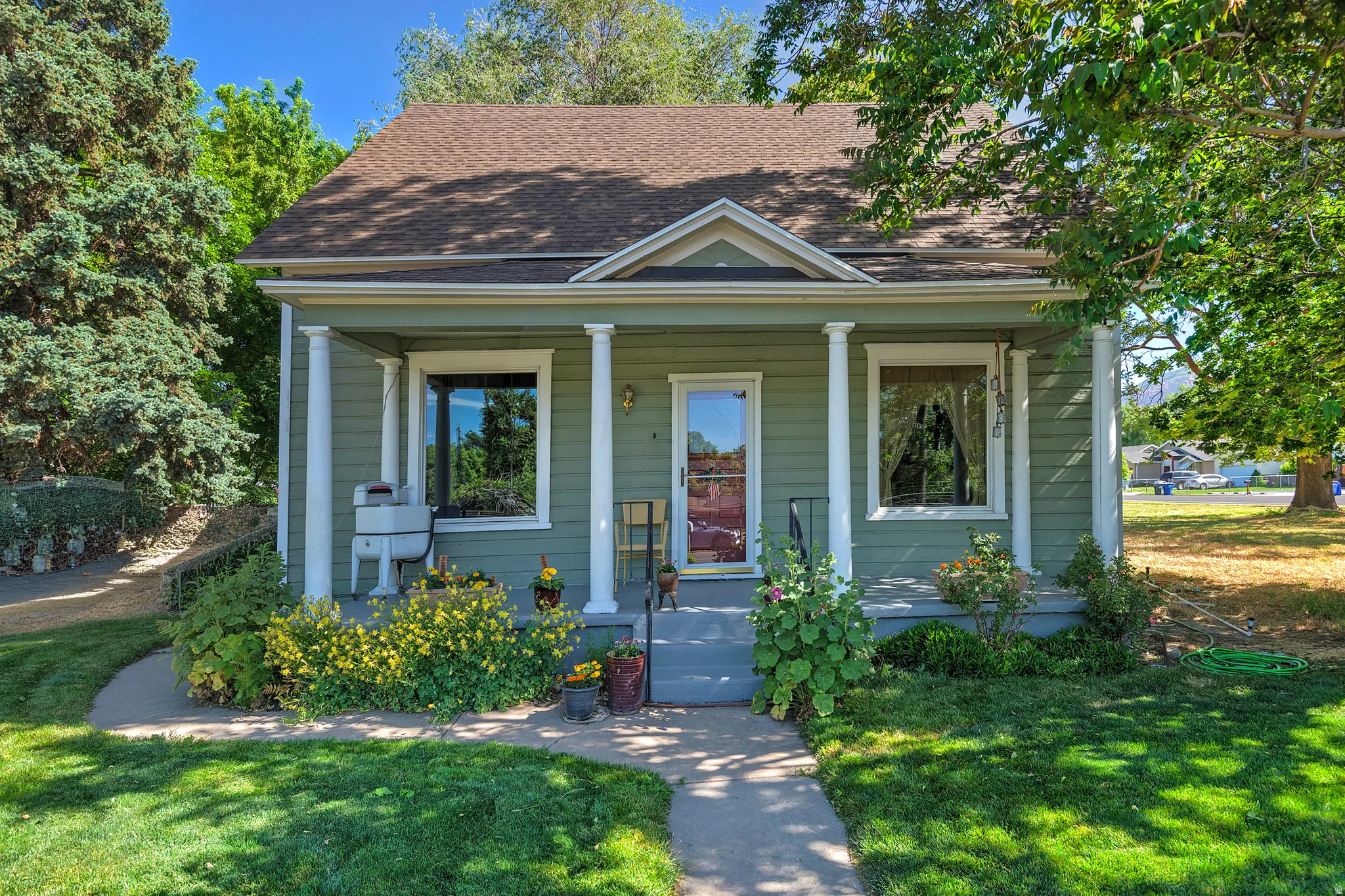 Green exterior with white trim featuring a covered porch with white columns, a dark shingle roof, and a mature tree providing shade
