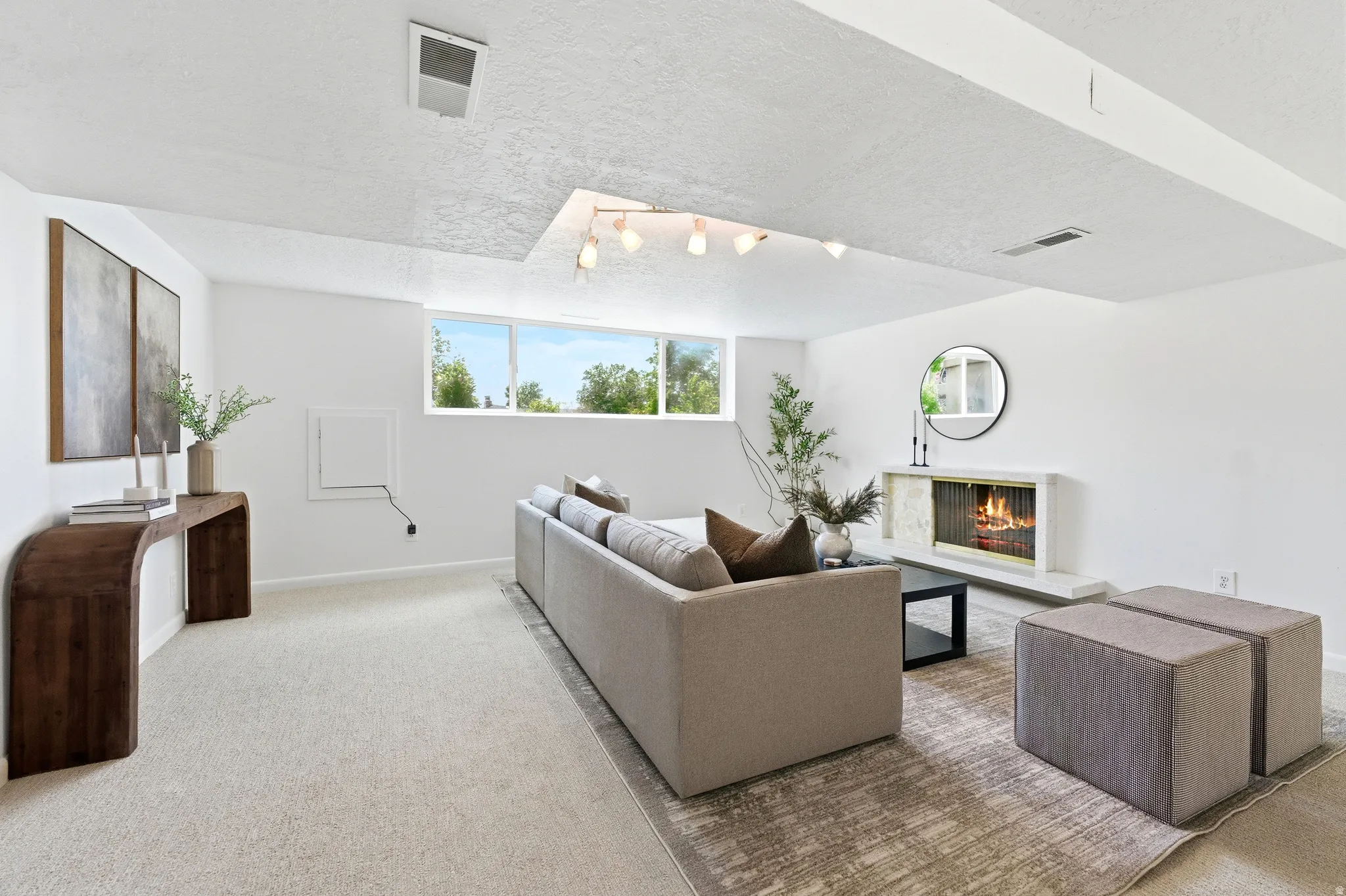 Spacious room featuring a fireplace with a light-toned surround, a built-in linear light fixture, a window providing natural light, and light-colored carpeting