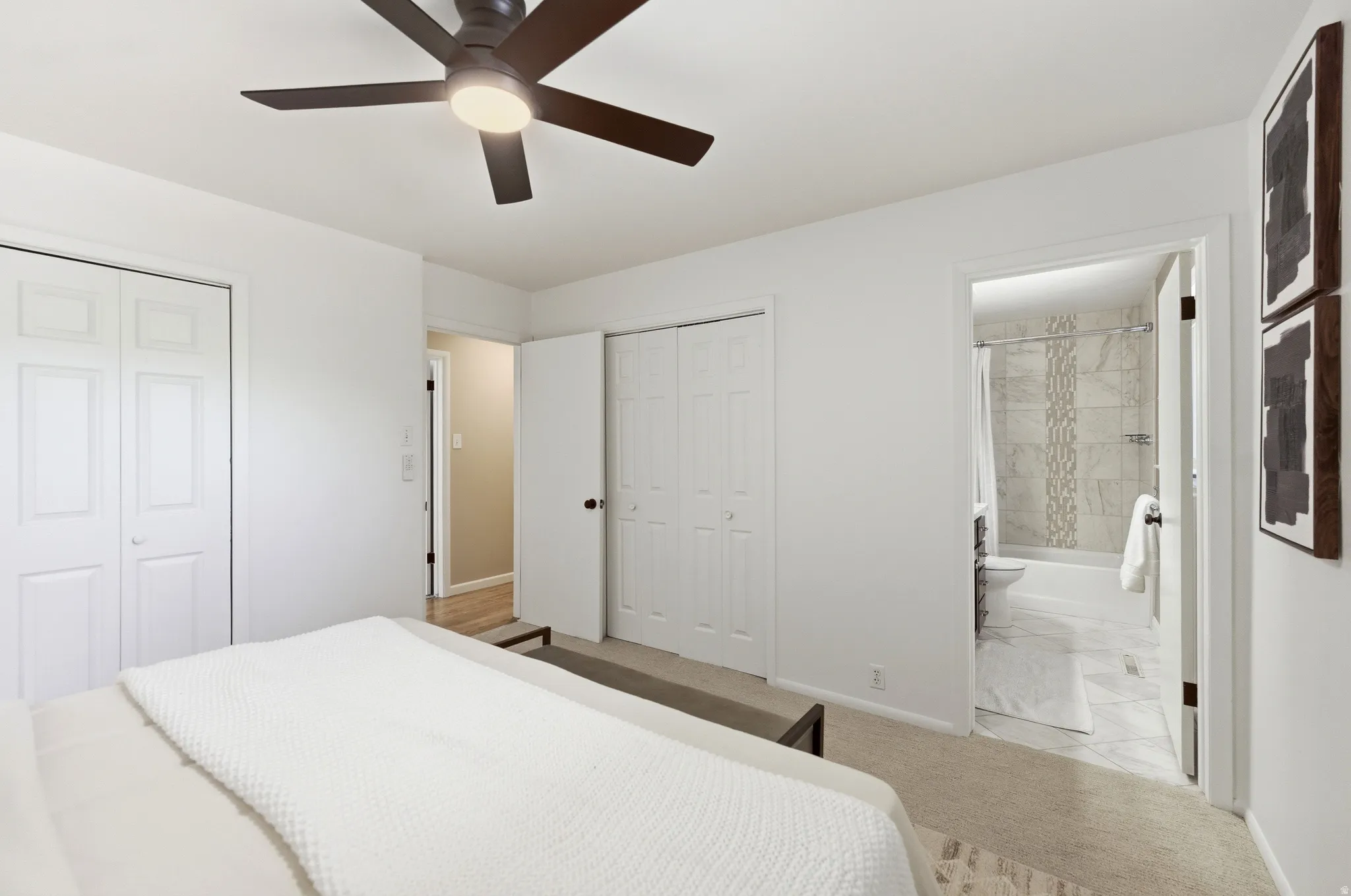 Neutral colored bedroom featuring a ceiling fan with integrated lighting, two bi-fold closet doors, and an en-suite bathroom with tiled flooring and a tiled shower surround