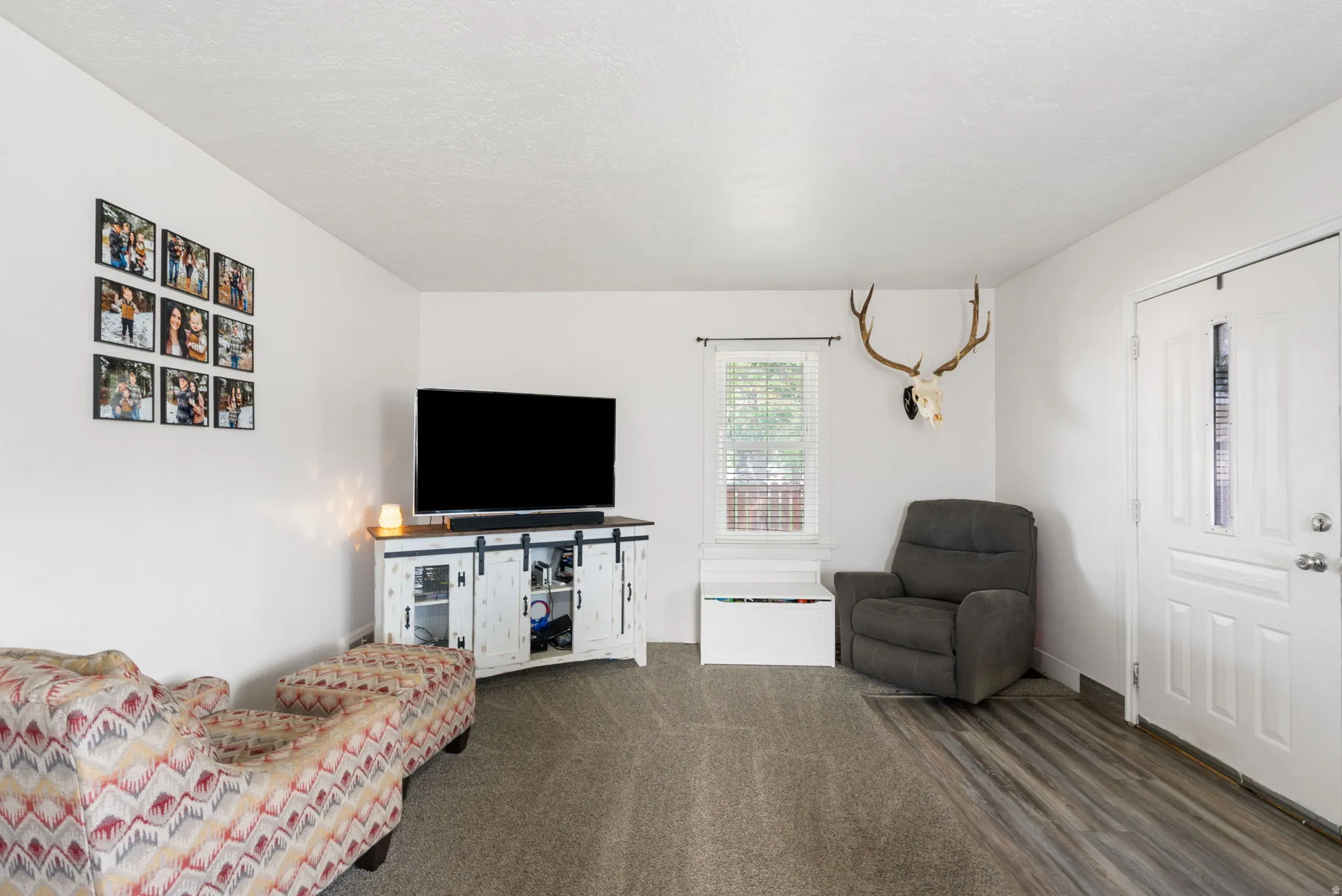 Living area featuring a textured gray carpet, wood-finish flooring, white walls, and a window with blinds