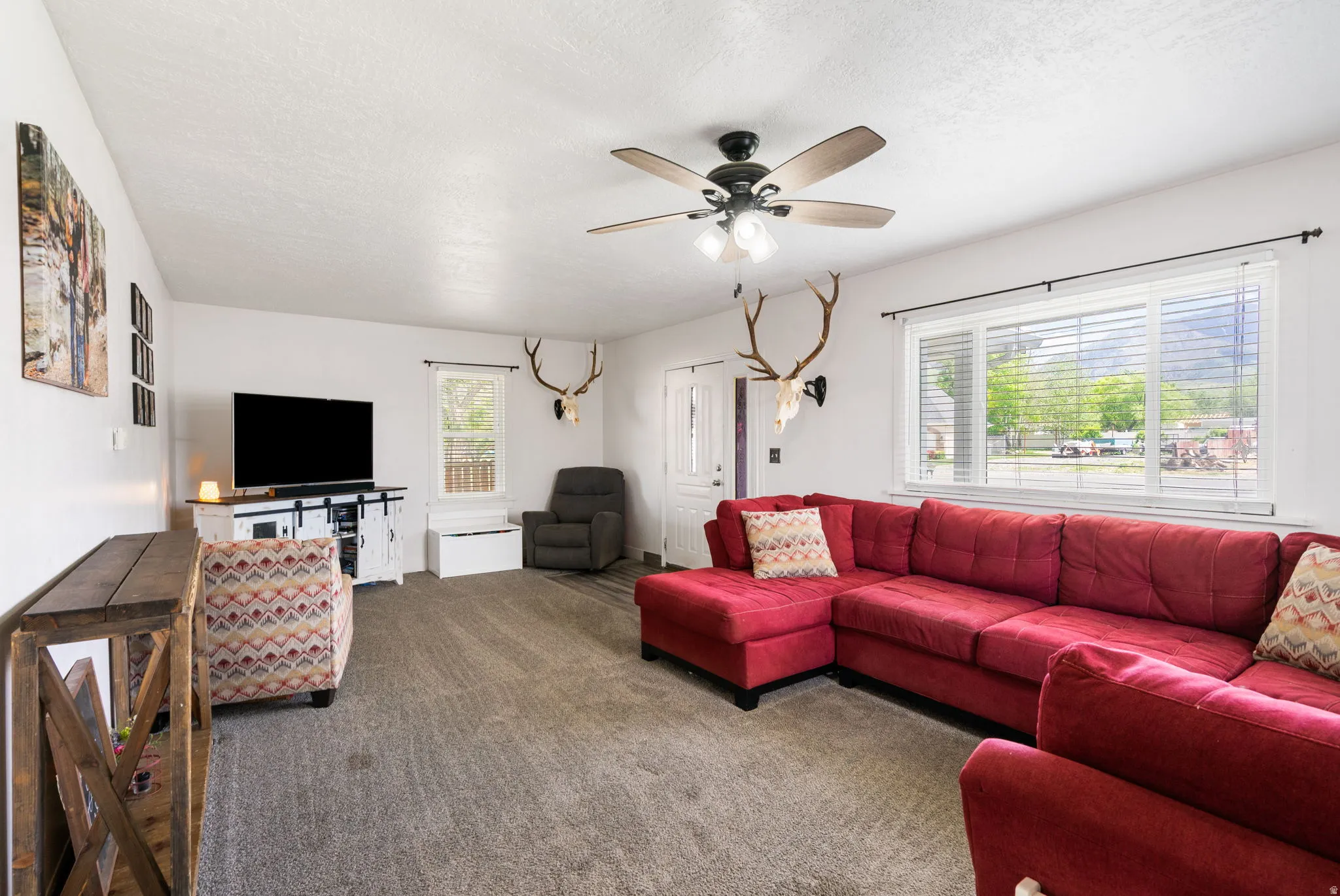 Spacious living area featuring neutral carpeting, a large picture window with horizontal blinds, a ceiling fan with integrated lighting, and a white paneled entry door