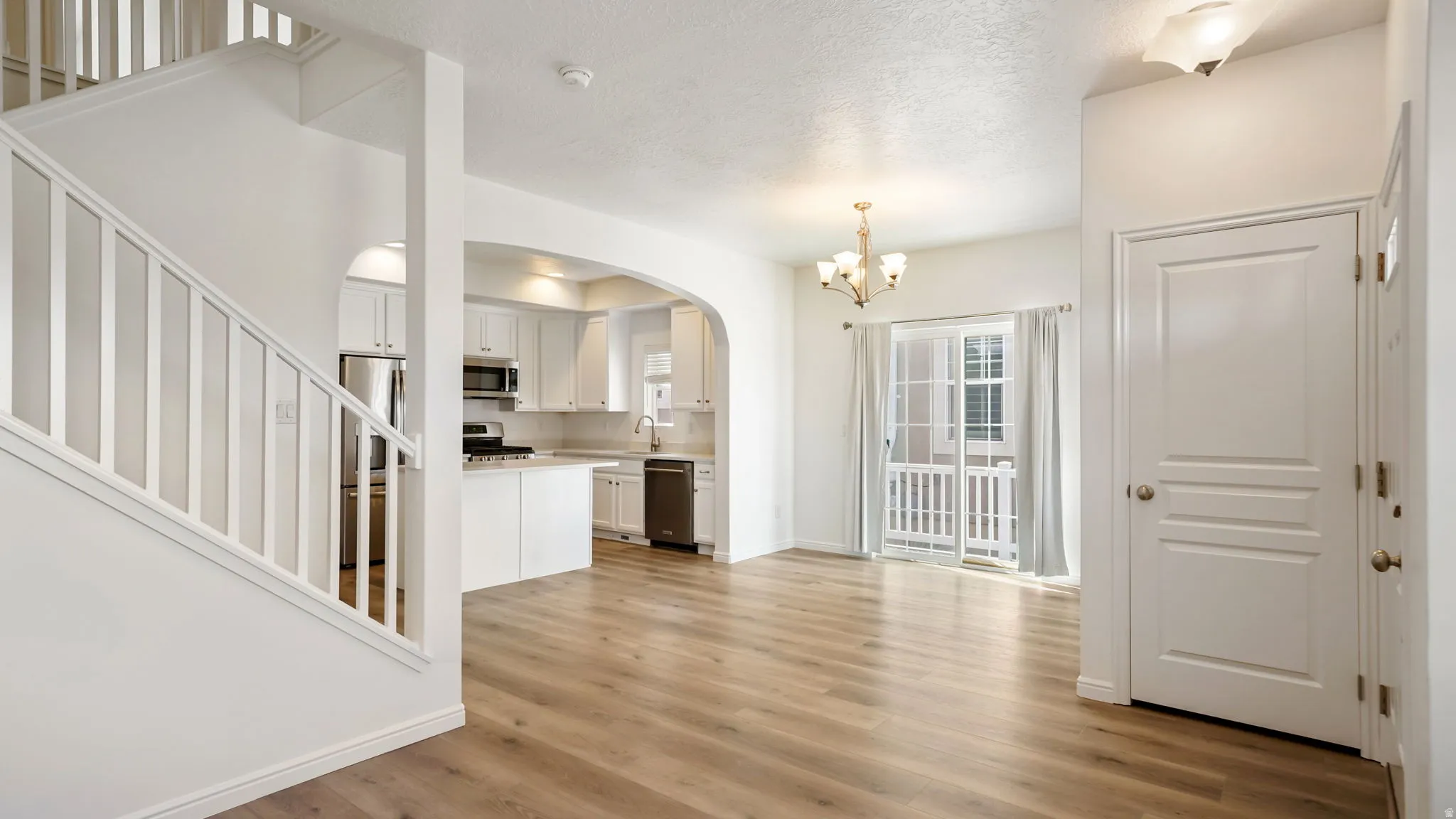 Open-concept living area featuring wood-finish flooring, a white kitchen with stainless steel appliances, an arched entryway, a white staircase, and a sliding glass door leading to an exterior balcony
