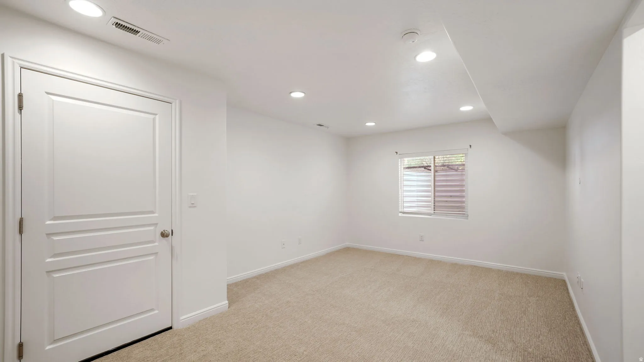 Bright room featuring recessed lighting, a neutral carpet, a white paneled door, and a single window with blinds