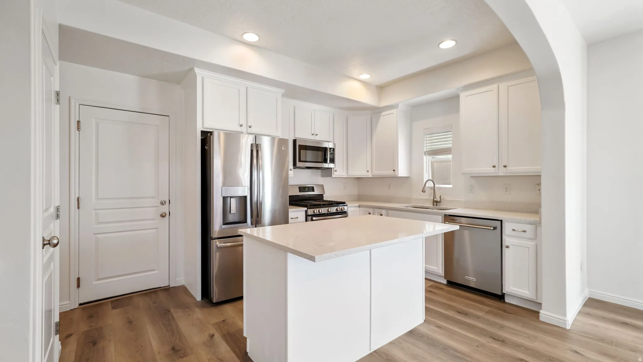 Modern kitchen featuring white cabinetry, stainless steel appliances, wood-finish flooring, a central island with a light-toned countertop, and recessed lighting