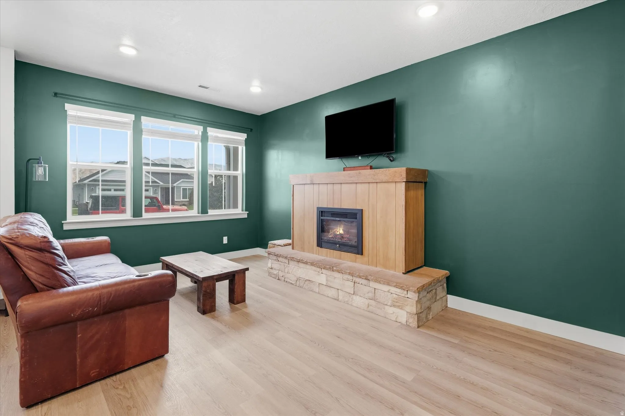 Living area featuring a floor-to-ceiling fireplace with a stone base and wood-finish surround, recessed lighting, wood-finish flooring, and a triple window assembly