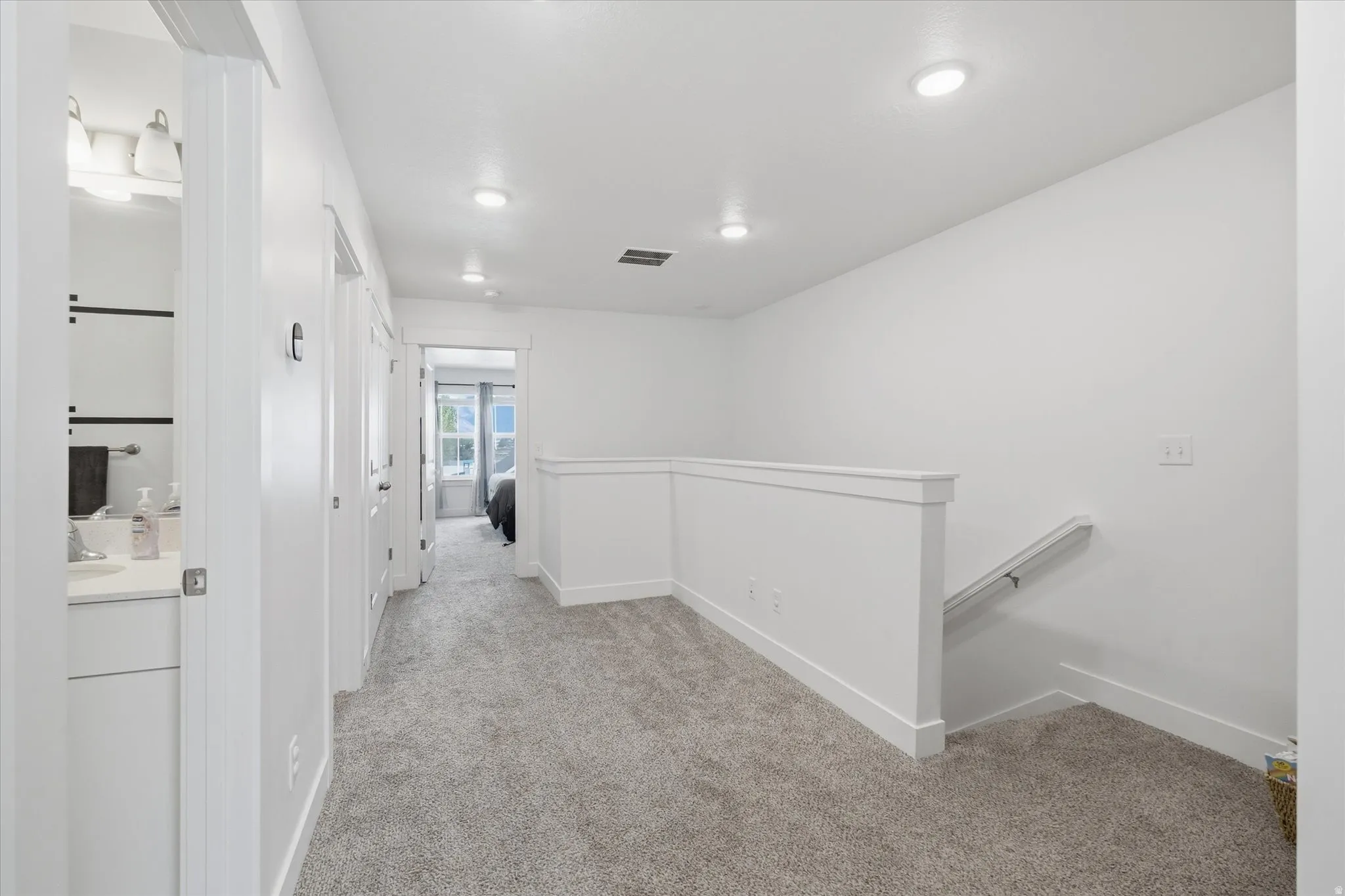Carpeted hallway with white walls and trim, featuring recessed ceiling lighting, a partial staircase with a handrail, and white paneled doors