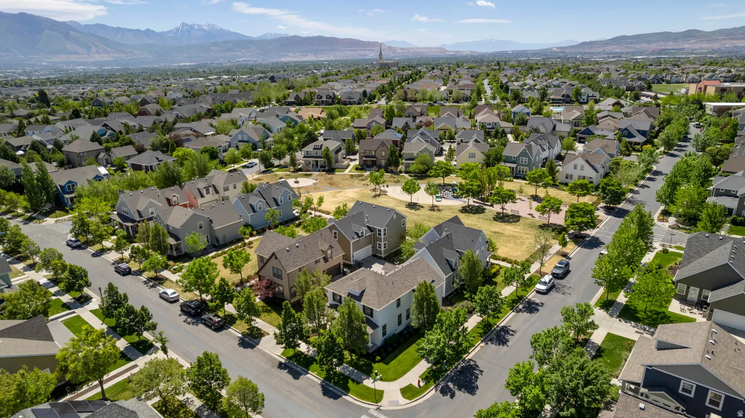 Aerial perspective showcasing a residential neighborhood with diverse architectural styles, tree-lined streets, a central green space, and distant mountain ranges