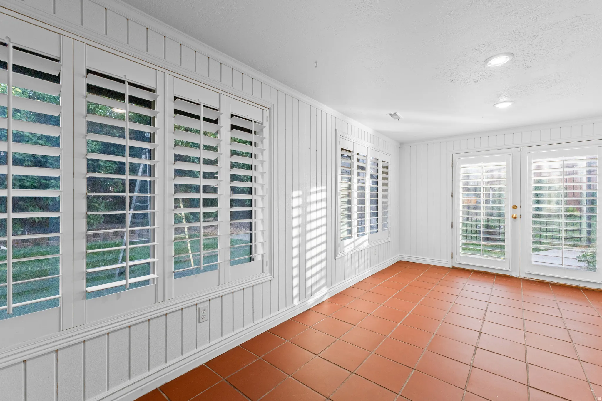 Spacious room featuring terra cotta tile flooring, white vertical wall paneling, multiple windows with plantation shutters, French doors, and recessed ceiling lighting