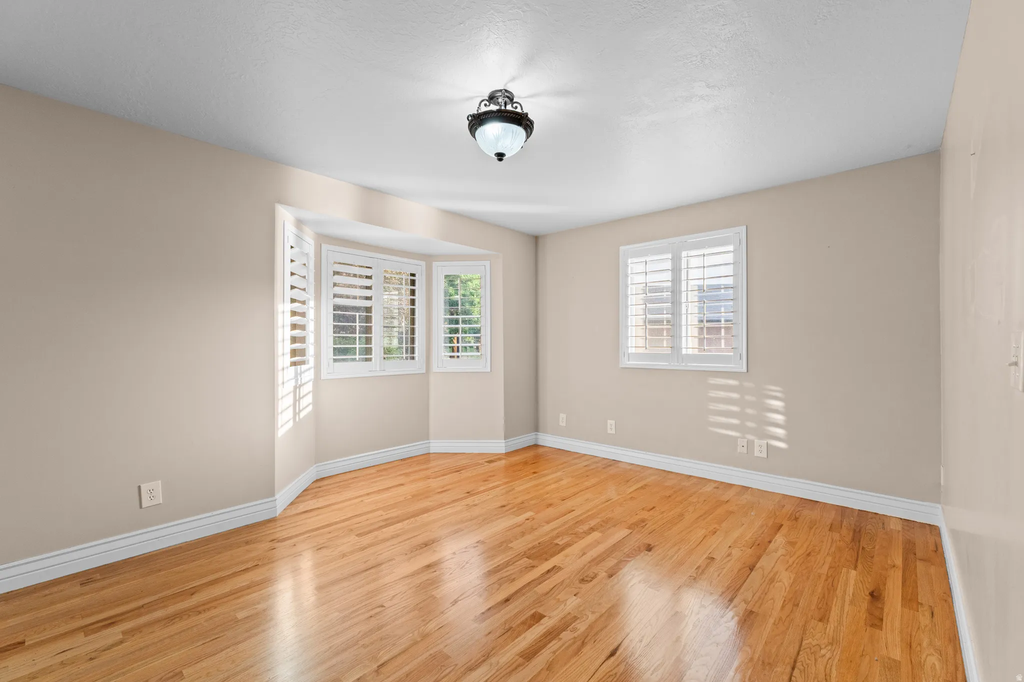 Bedroom featuring wood-finish flooring, a bay window with white plantation shutters, a standard window with white plantation shutters, neutral wall tones, and a ceiling-mounted light fixture