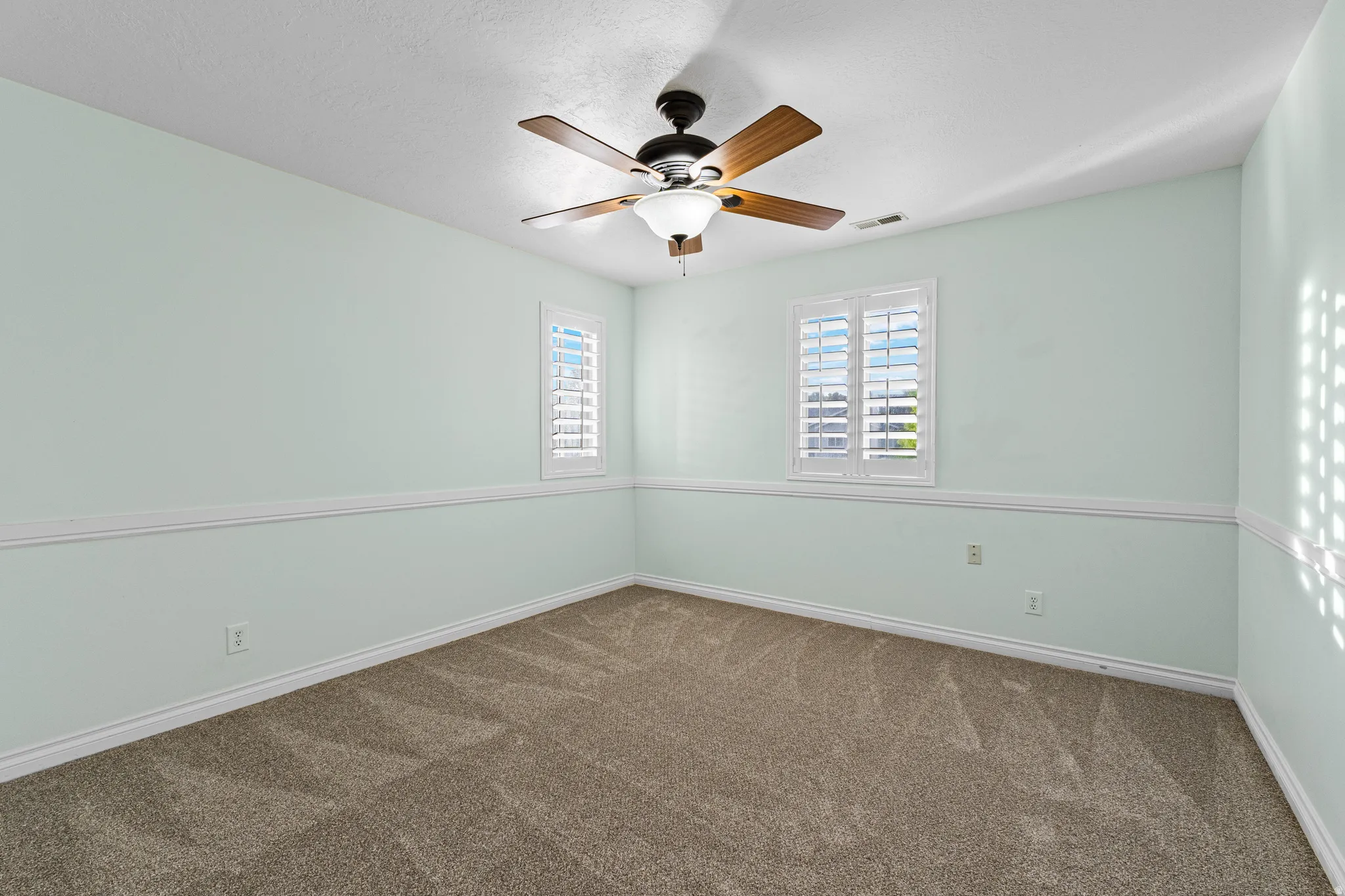 Carpeted room featuring a ceiling fan with light fixture, two windows with white plantation shutters, and decorative wall molding